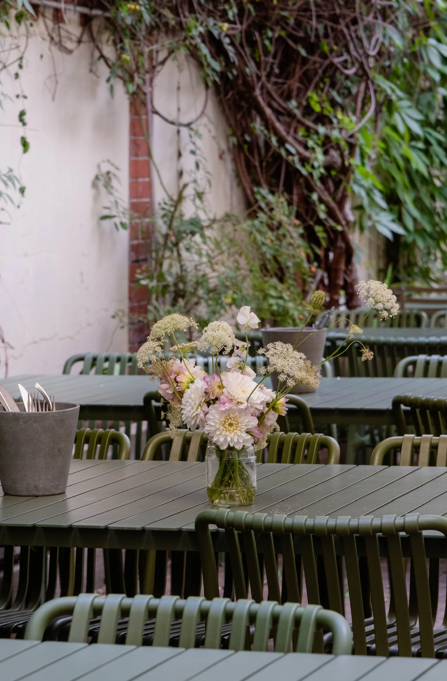 Outdoor patio with a round table, a glass jar with a bouquet of light pink and white flowers as a centerpiece, and green chairs. In the background, there is a wall with climbing vines and a large twisted vine.