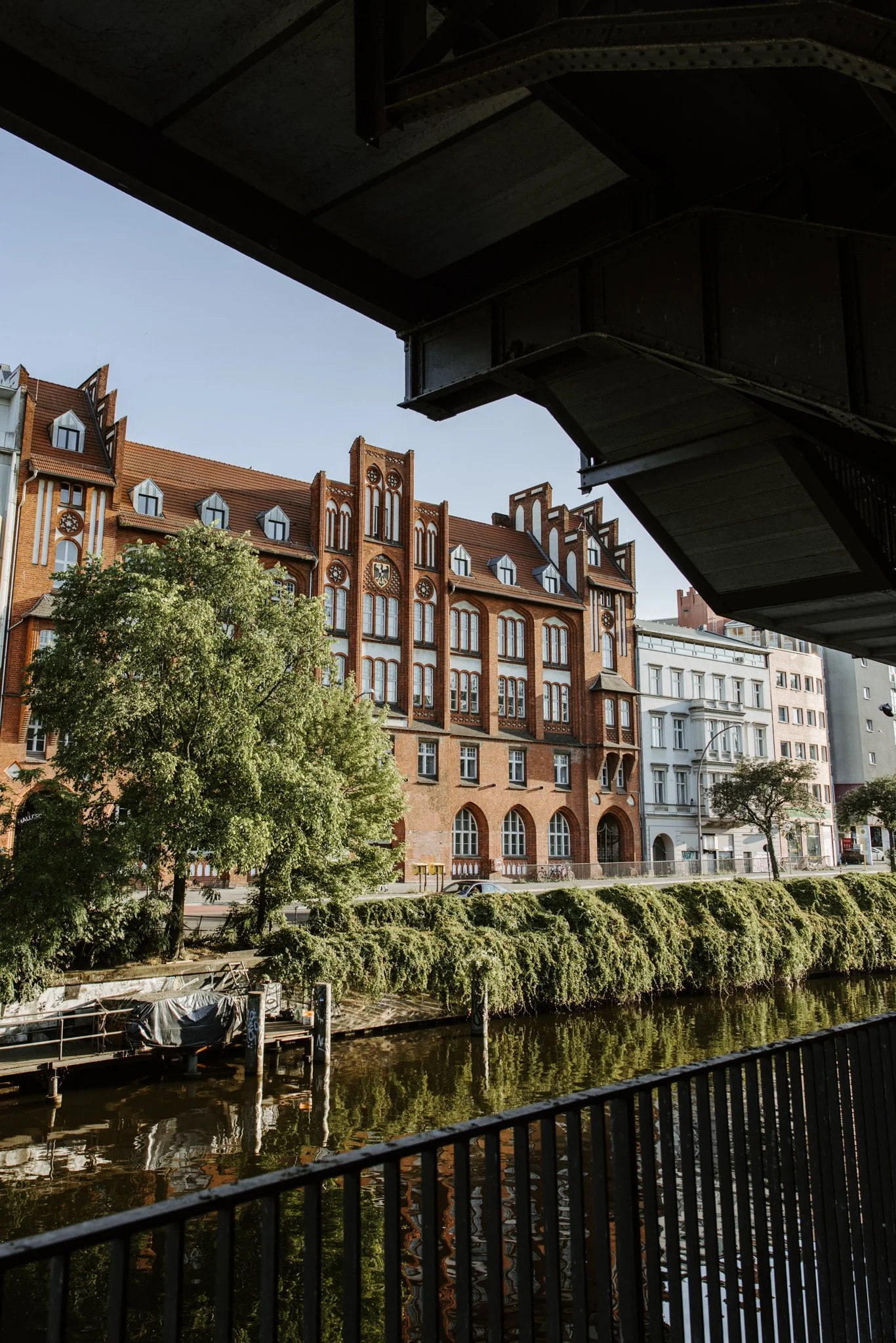 View of historic red brick building with pointed gables and large windows along a riverfront, seen from beneath a bridge over the water with trees and a boat dock in the foreground.