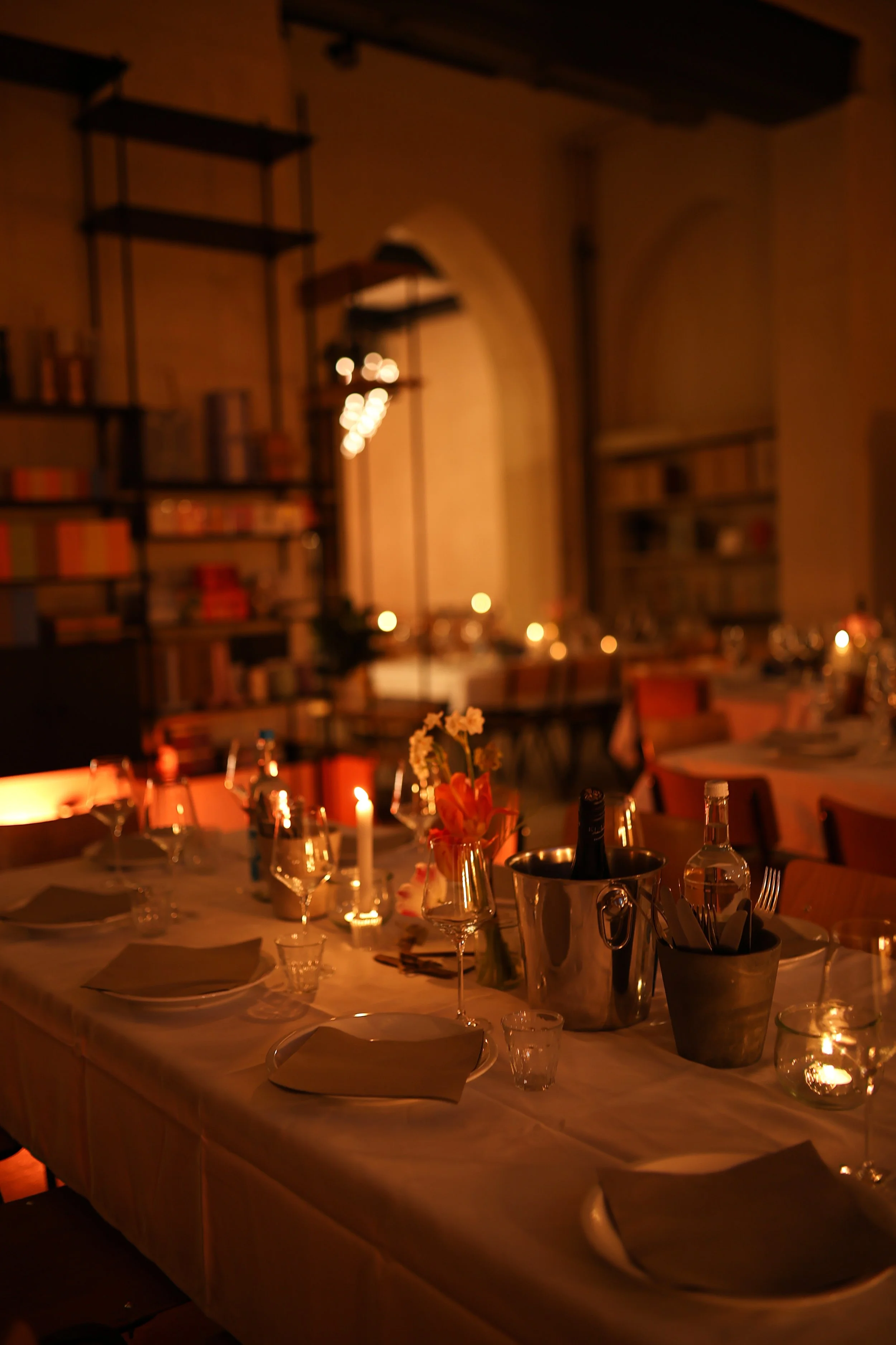 A warmly lit banquet table set for dinner with wine glasses, candles, flowers, and utensils in a cozy, book-filled restaurant or library setting.