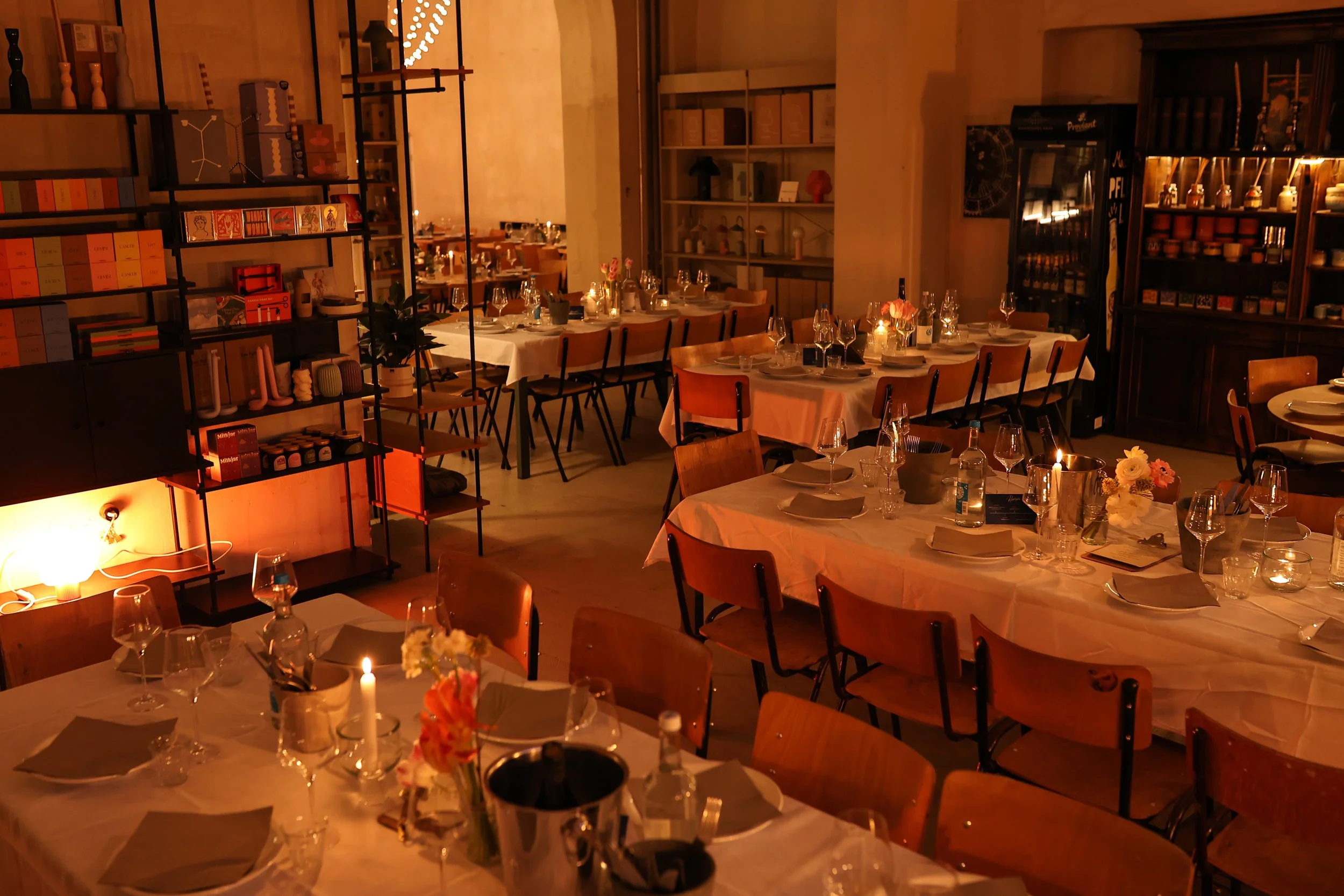 A cozy, warmly lit restaurant or event space with tables set for dining, decorated with candles and flowers, and surrounded by shelves and wooden furniture.