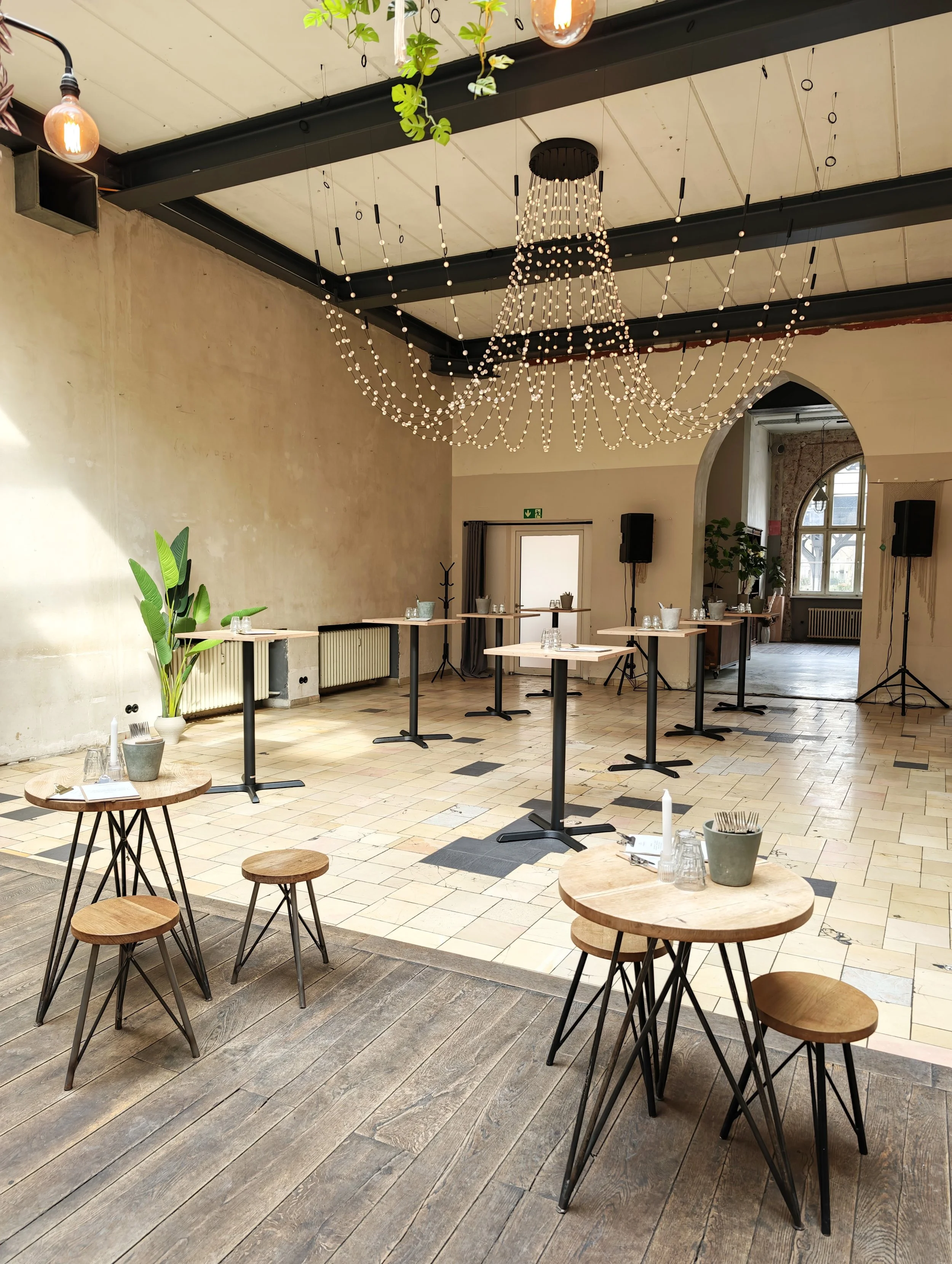 Interior of a modern restaurant or event space with high ceilings, string lights, wooden tables, and chairs. Decor includes a large hanging chandelier with lights, a potted plant, and some small tables with glassware and menus.