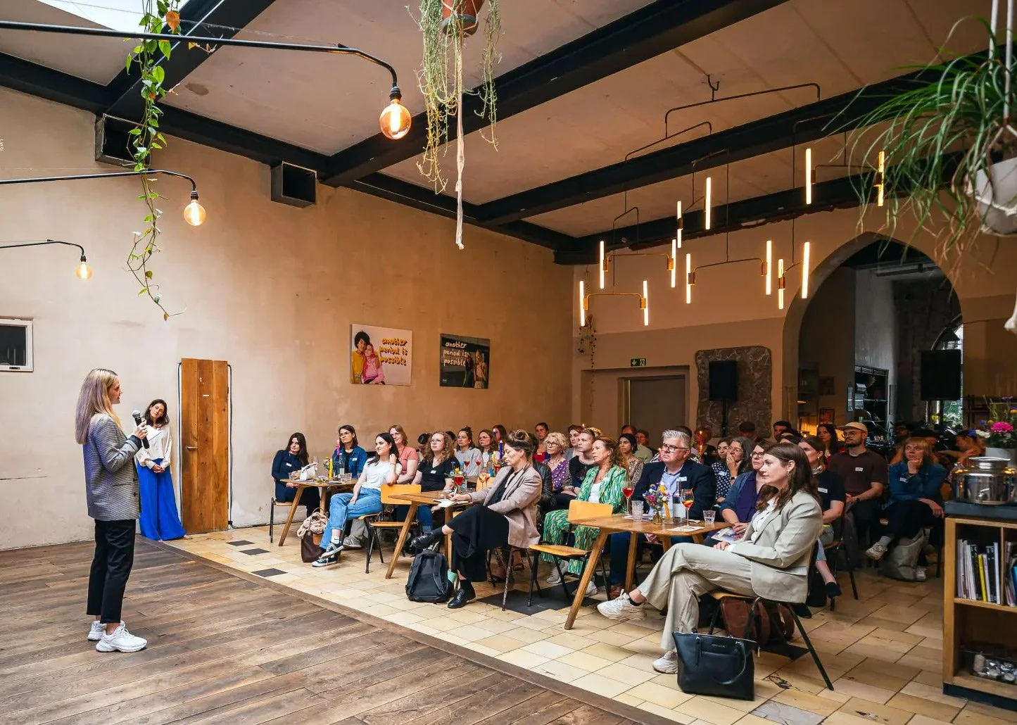A woman stands on a stage speaking into a microphone to an audience in a large, modern room with hanging light fixtures, plants, and bookshelves.