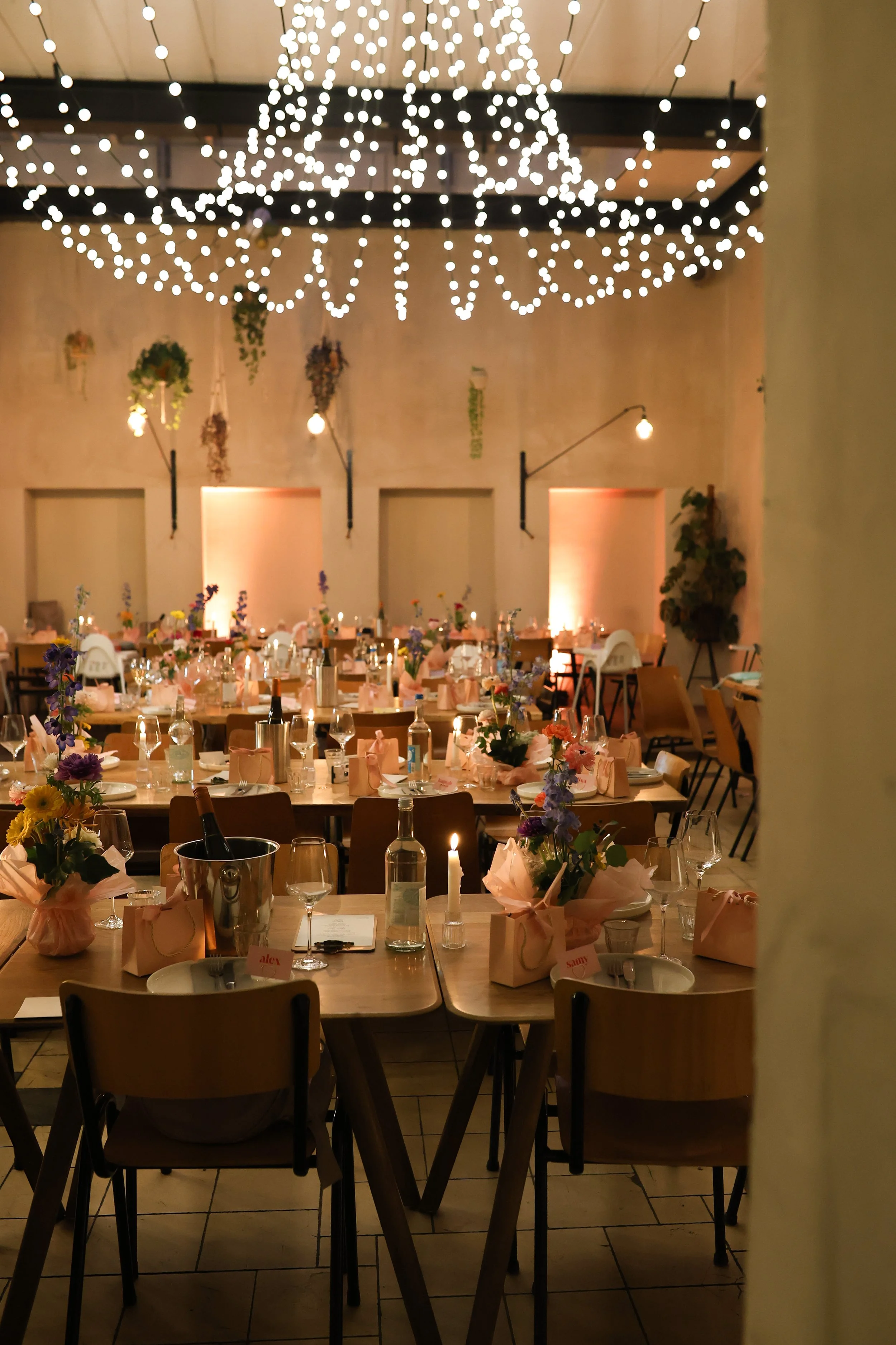 A banquet hall decorated for a celebration with tables set for guests, adorned with flowers, candles, and place settings; a large chandelier hangs from the ceiling with string lights creating a festive atmosphere.