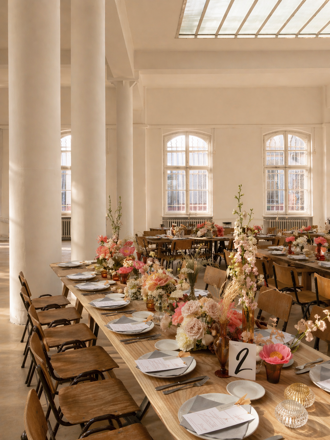 Long dining table with pink, white, and cream floral centerpieces set for a wedding or event in a bright, spacious room with large windows and white columns.