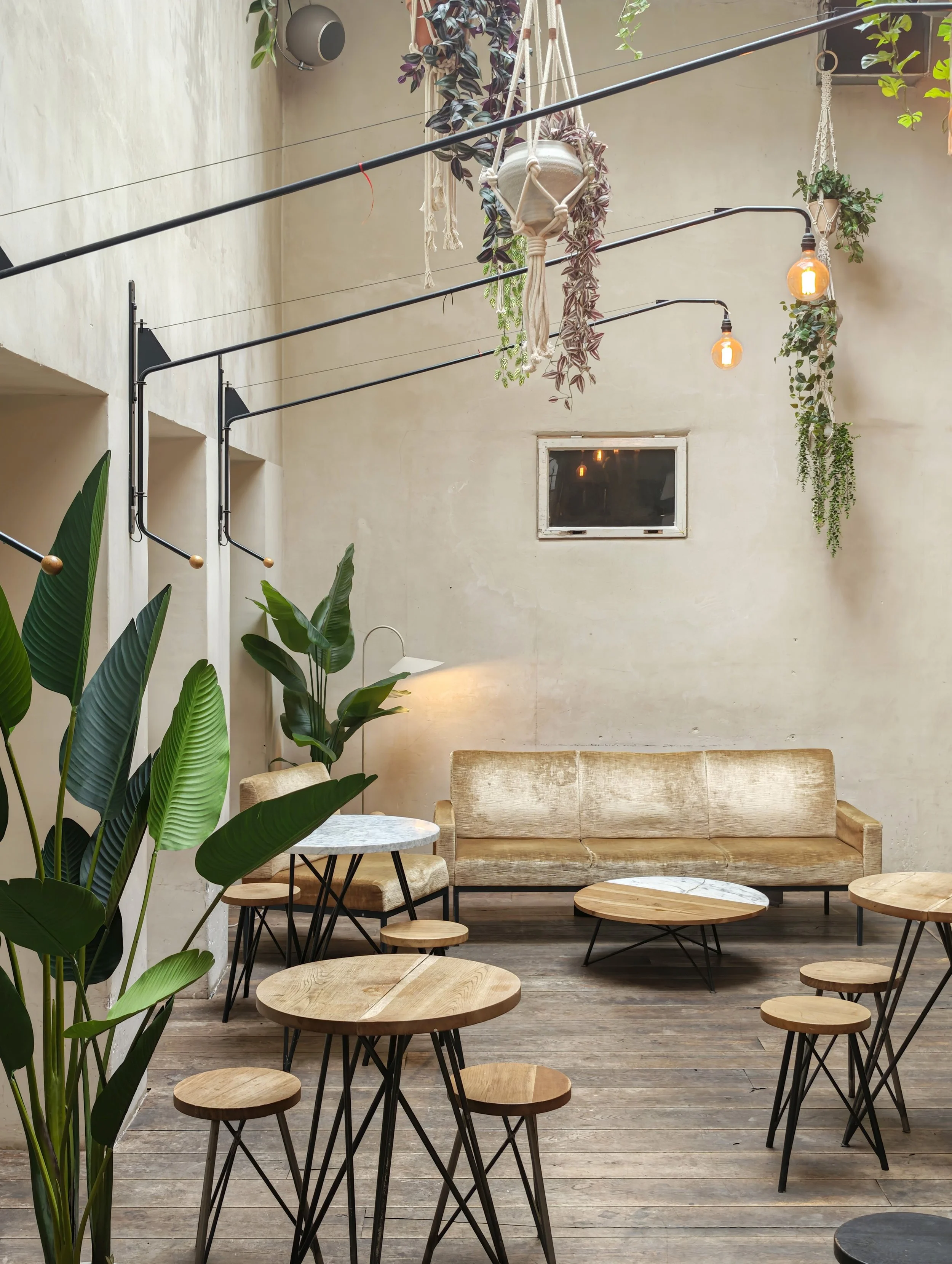 Interior of a stylish cafe or lounge with wooden tables, round stools, a beige velvet sofa, and lush green plants. Hanging lightbulbs and macramé planters add to the cozy, modern decor.