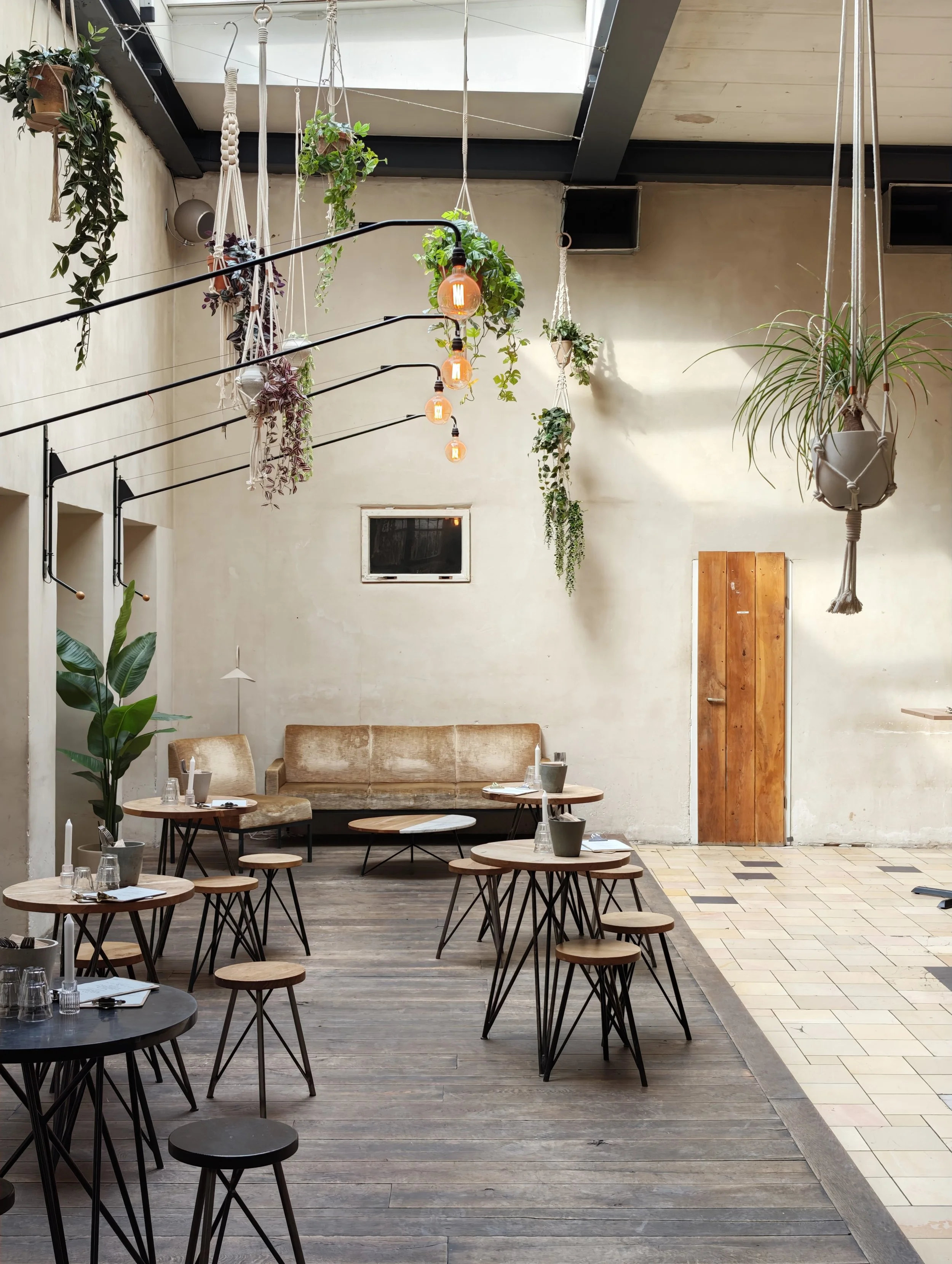 Interior of a modern café with tables, stools, hanging plants, and hanging light bulbs, with a mix of wood and tile flooring and a high ceiling with a skylight.