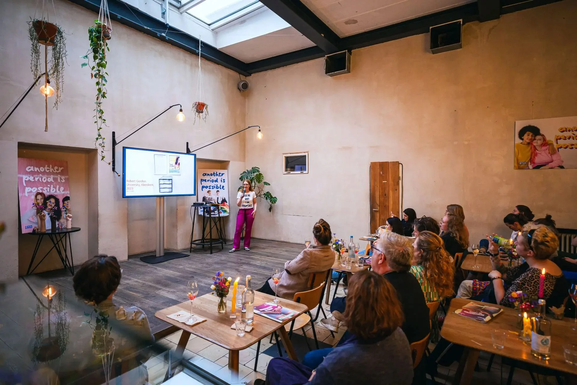 A woman giving a presentation to an audience in a well-lit room with a high ceiling and a skylight. The room has hanging plants, wall art, and a large screen displaying a slide.