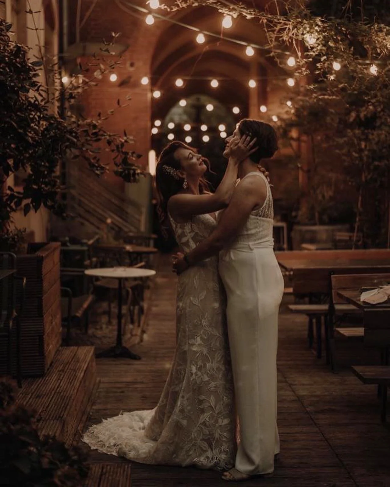A romantic moment between two women in wedding attire, holding each other in a warmly lit, outdoor setting at night with string lights overhead.