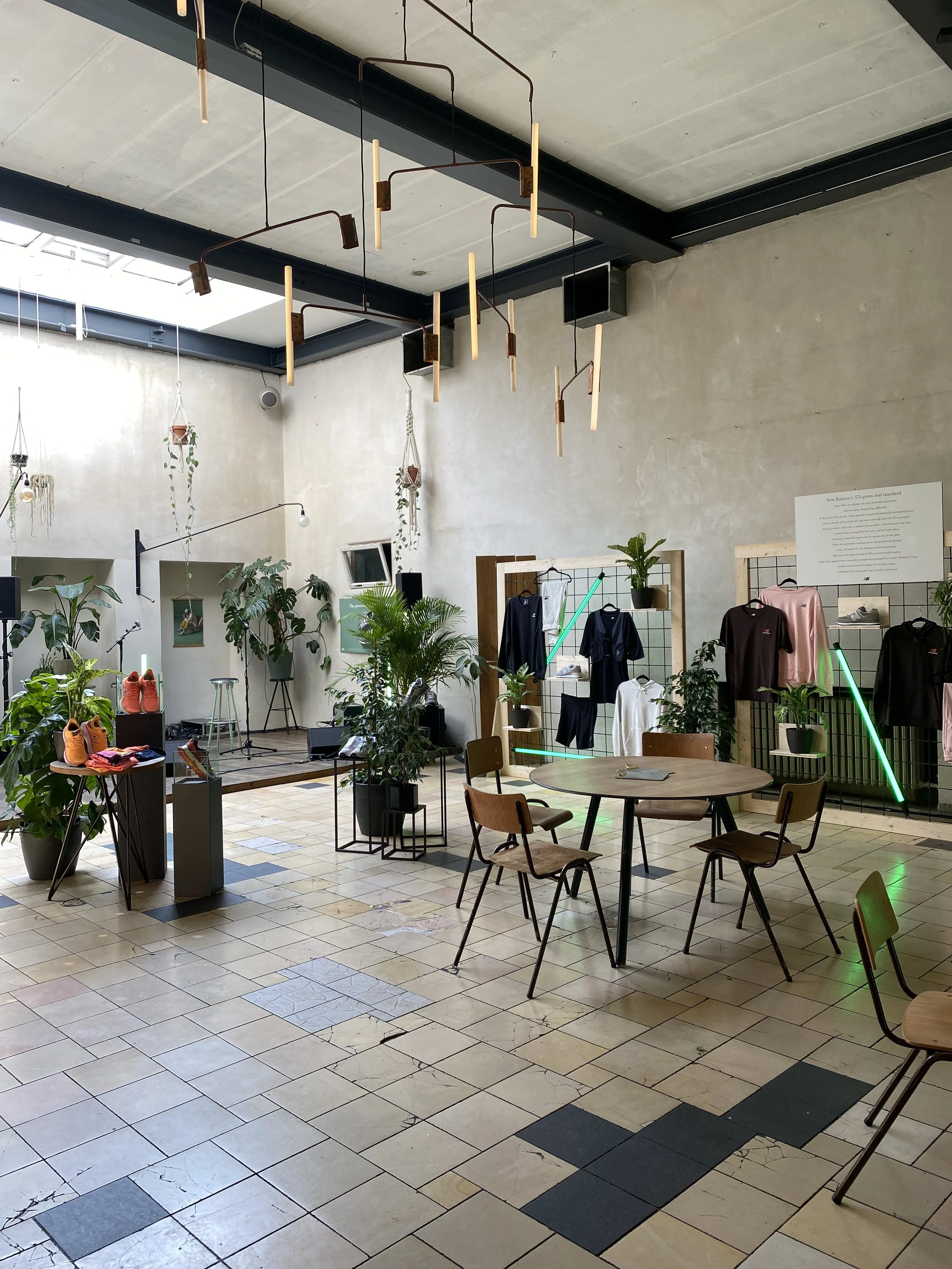 Interior of a modern retail or cafe space with plants, tables, chairs, clothing displays, and lighting fixtures on the ceiling, featuring a mix of industrial and natural decor.
