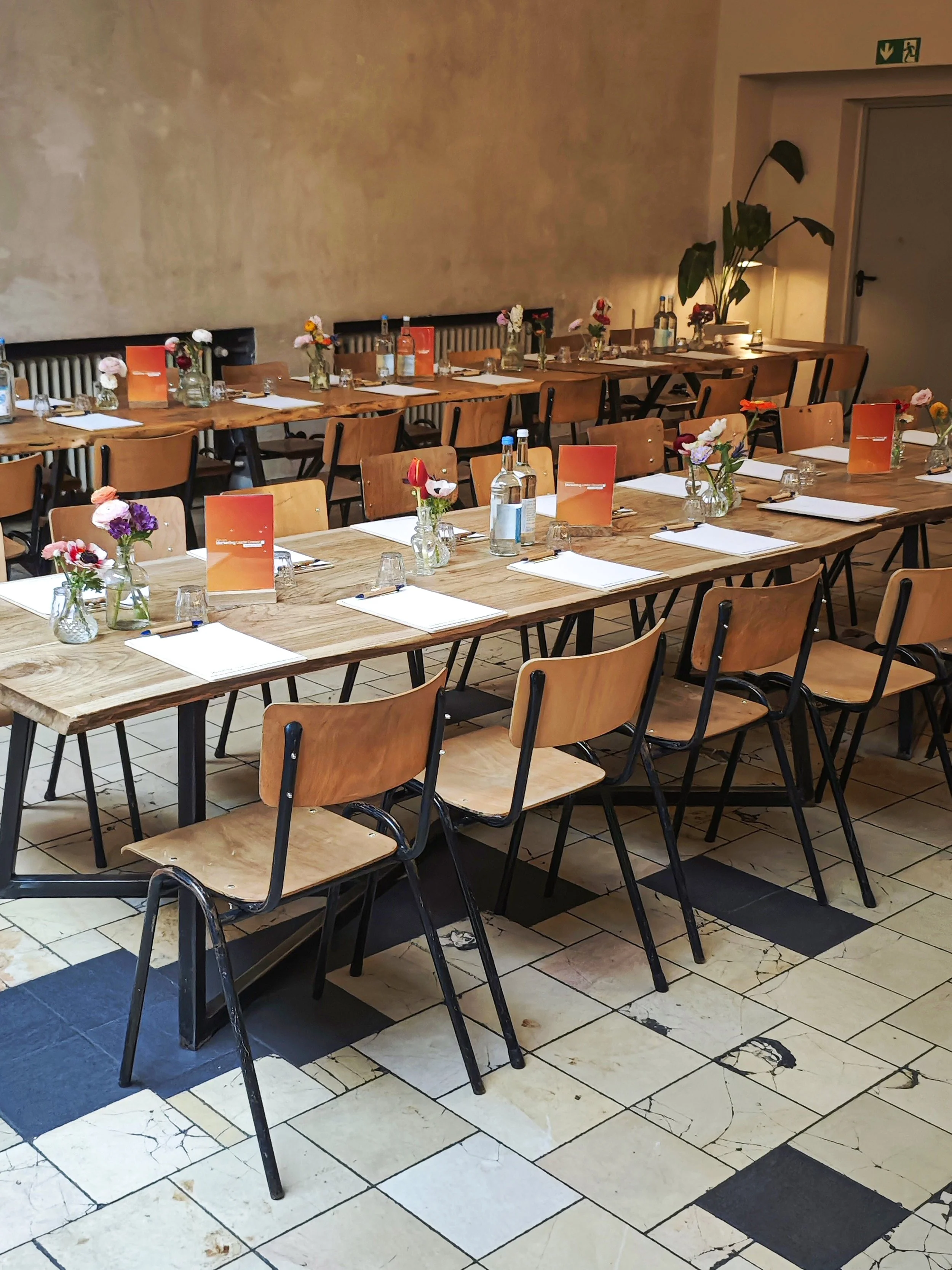A restaurant or cafe interior with a long wooden table set with white napkins, glasses, bottles of water, small vases with flowers, and orange menus. There are wooden chairs around the table, a tall plant in the background, and a wall with a beige te