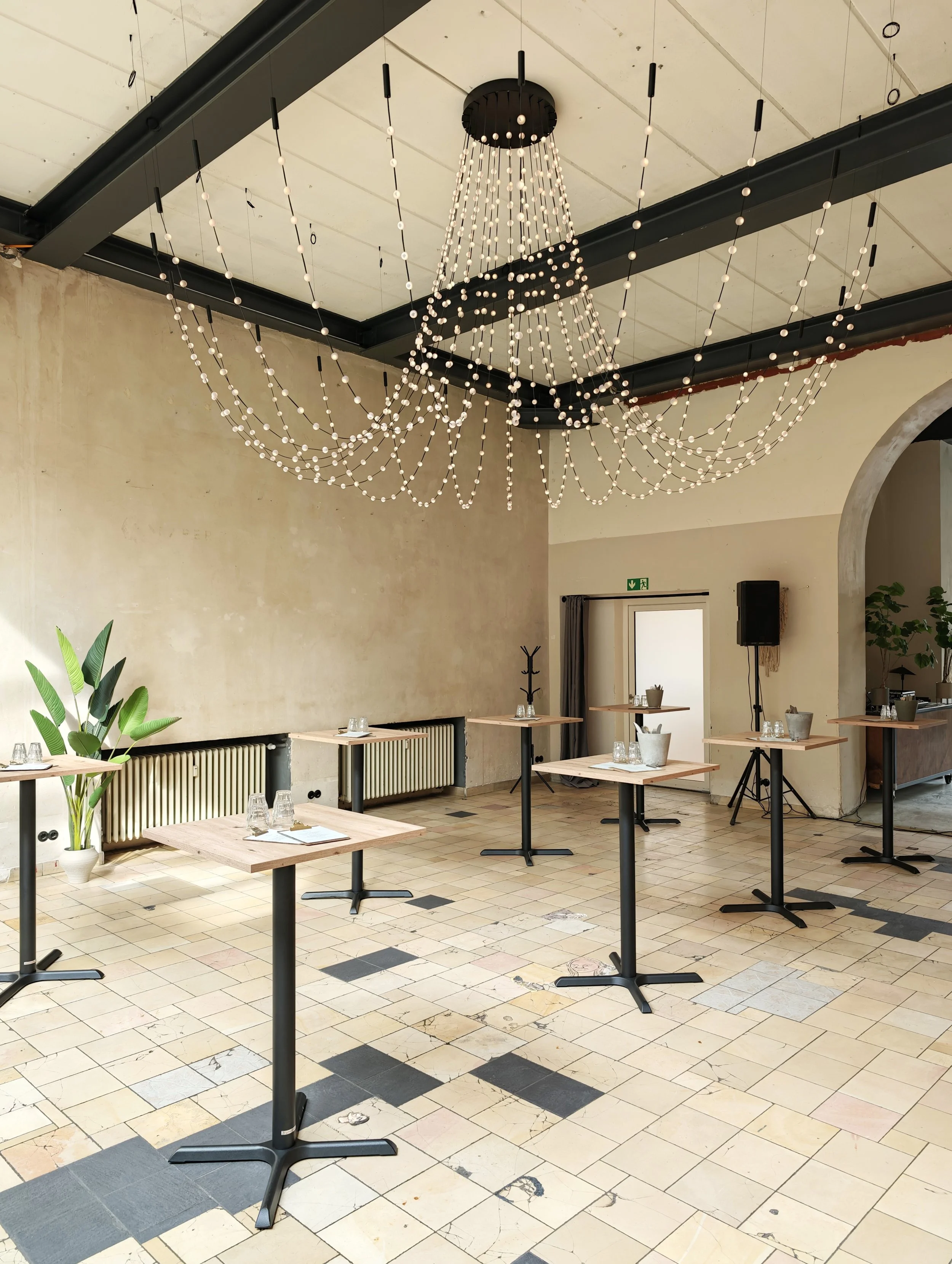 Empty indoor dining area with tables set with glasses and utensils, decorated with large modern chandelier, potted plants, and a textured tile floor.