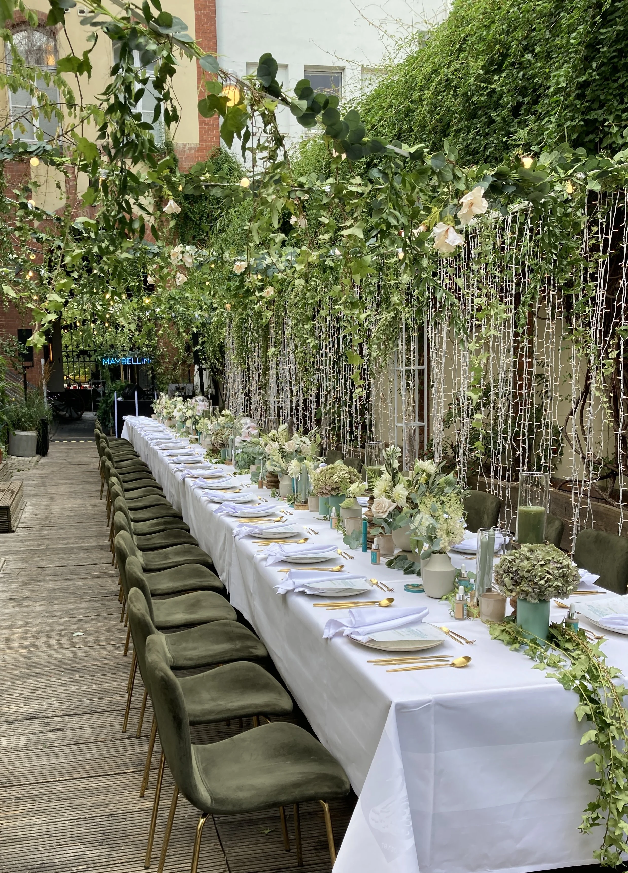 Long table set for a celebration with white tablecloths, floral centerpieces, and gold cutlery, outdoors on a wooden deck, decorated with hanging string lights and greenery overhead.