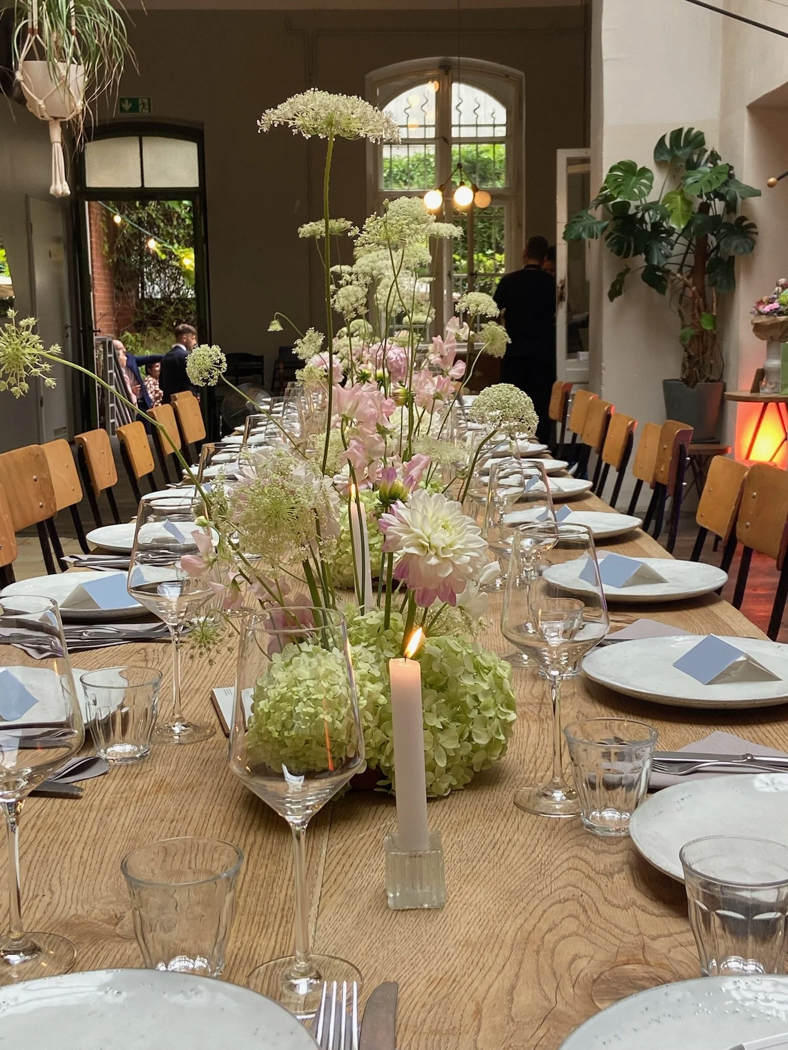 A long wooden dining table set for a formal event with white plates, silverware, wine glasses, and a white candle centerpiece, decorated with white and pink flowers.