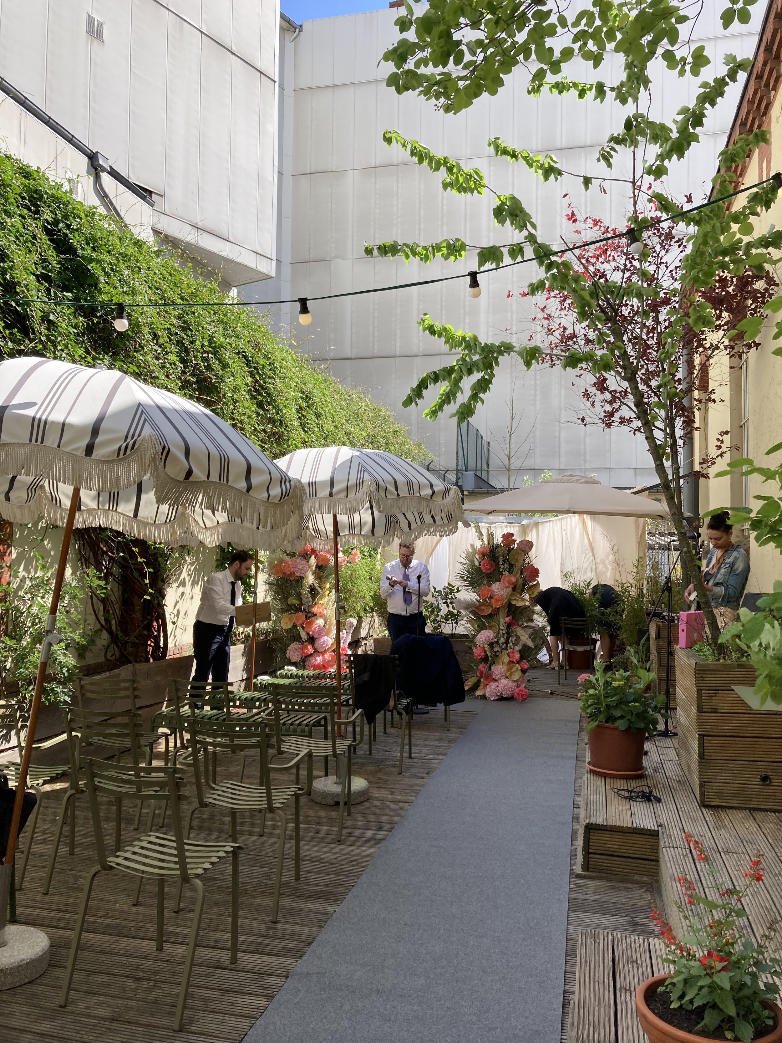 Outdoor courtyard setup for an event with chairs, umbrellas, potted plants, floral arrangements, and people preparing for a gathering under string lights and trees.
