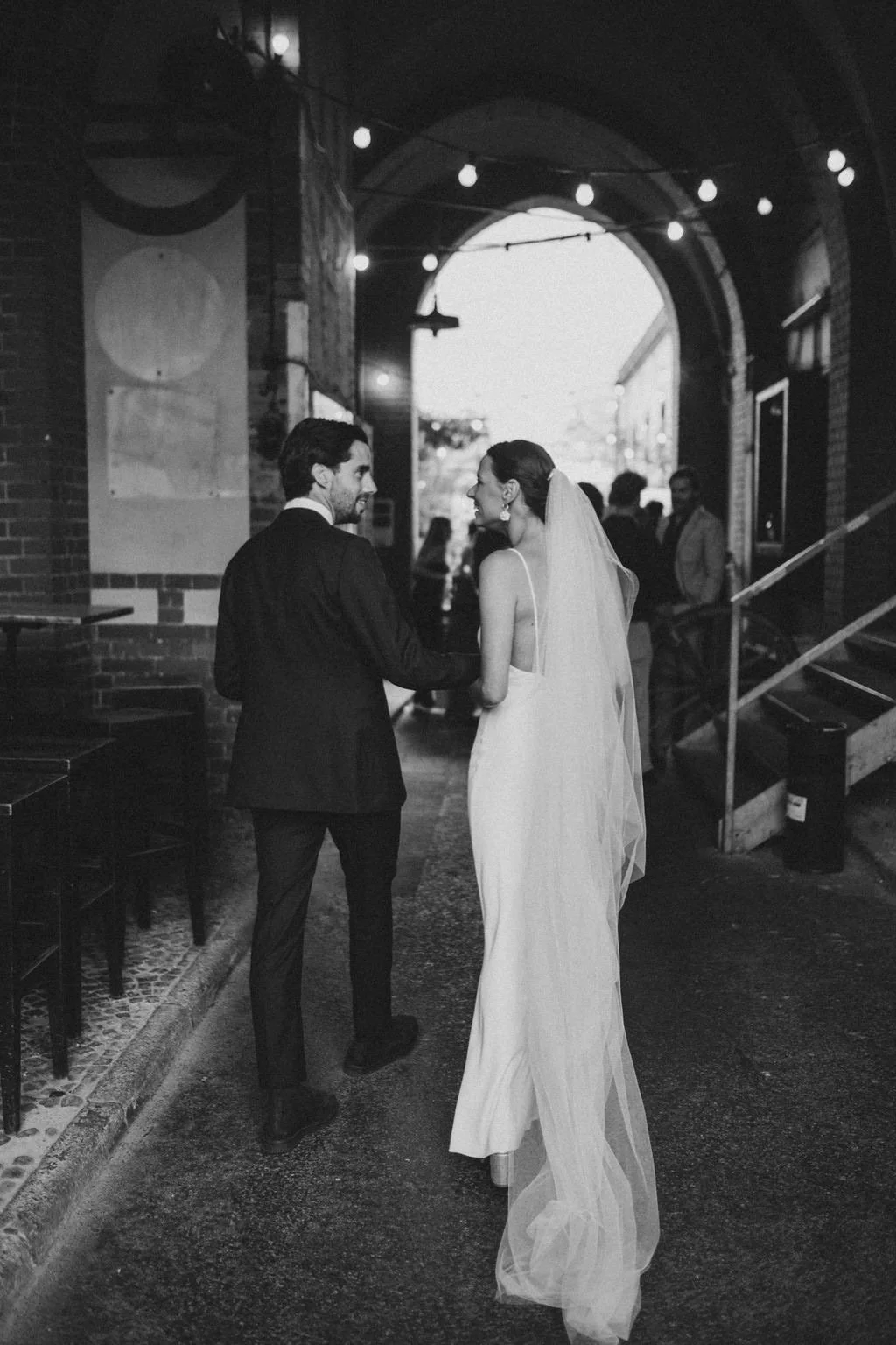 A black-and-white photo of a bride and groom holding hands, looking at each other beneath an arched entrance with string lights. The bride is in a long white wedding dress with a veil, and the groom is in a dark suit. There are a few people in the ba