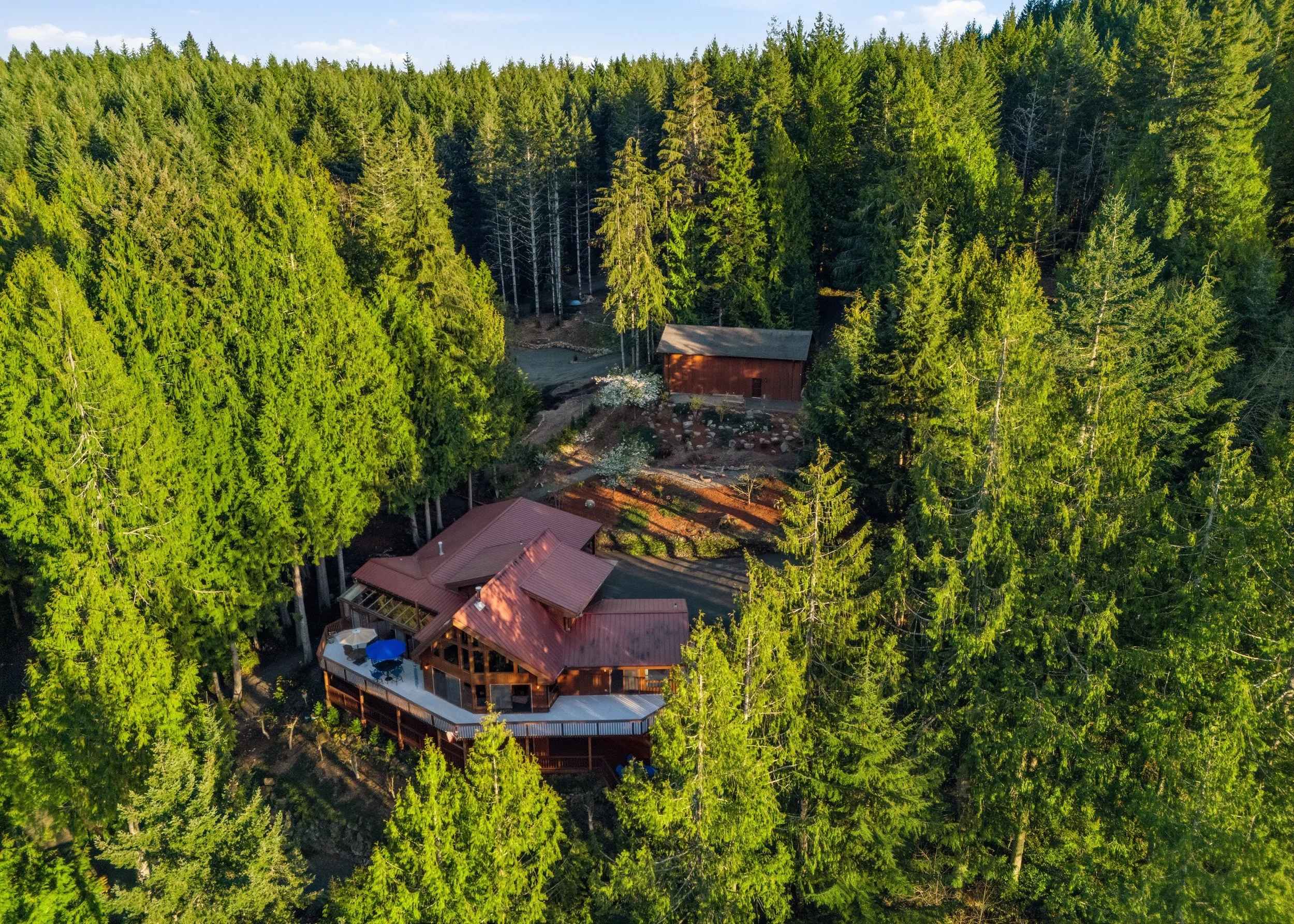 Aerial view of a house with a red roof and a spacious deck, surrounded by dense green trees in a forested area.