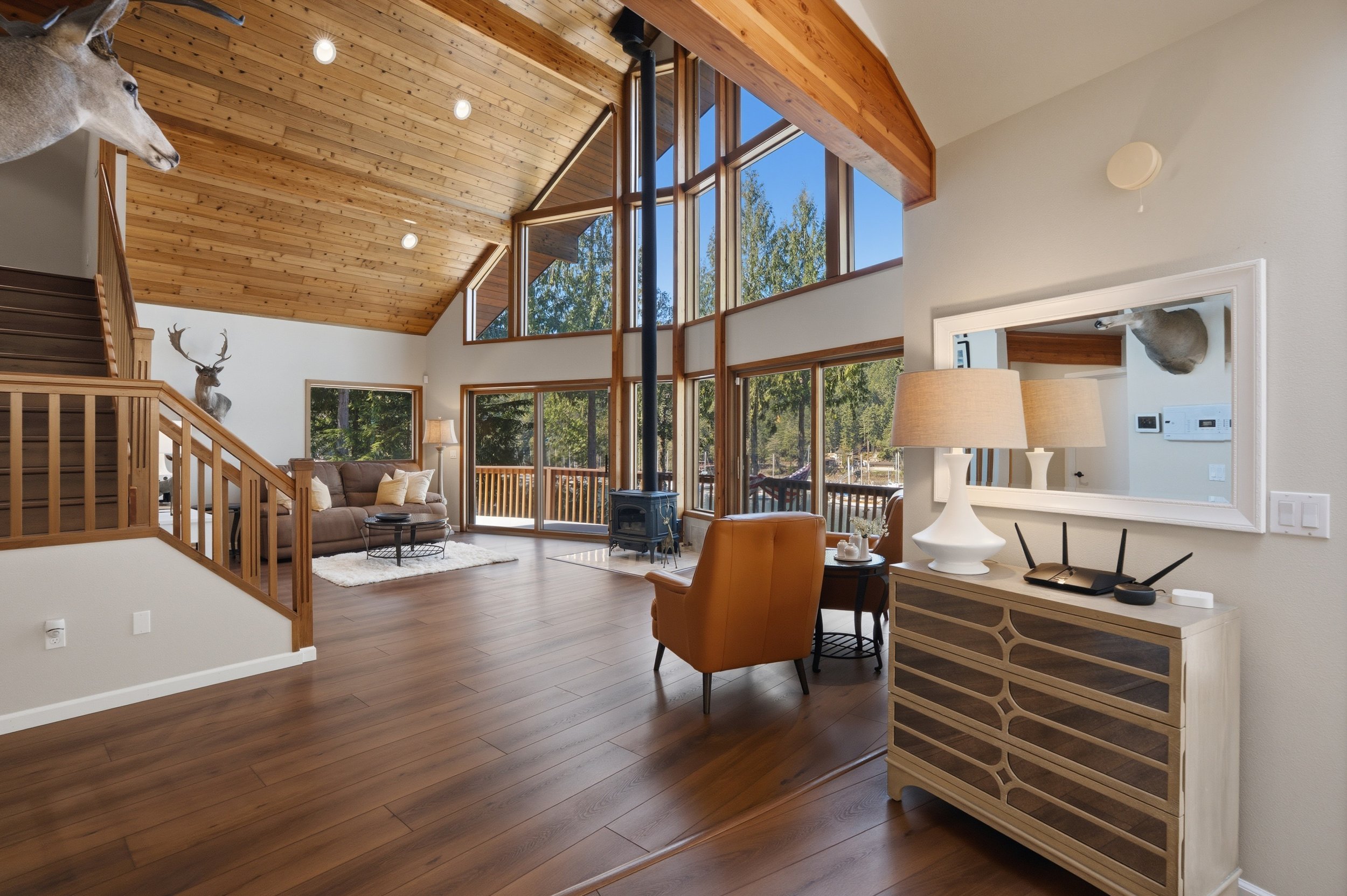 Spacious living room with large floor-to-ceiling windows, wood-paneled ceiling, wooden staircase, comfortable sofa, leather armchair, decorative deer mounts, and wood stove.