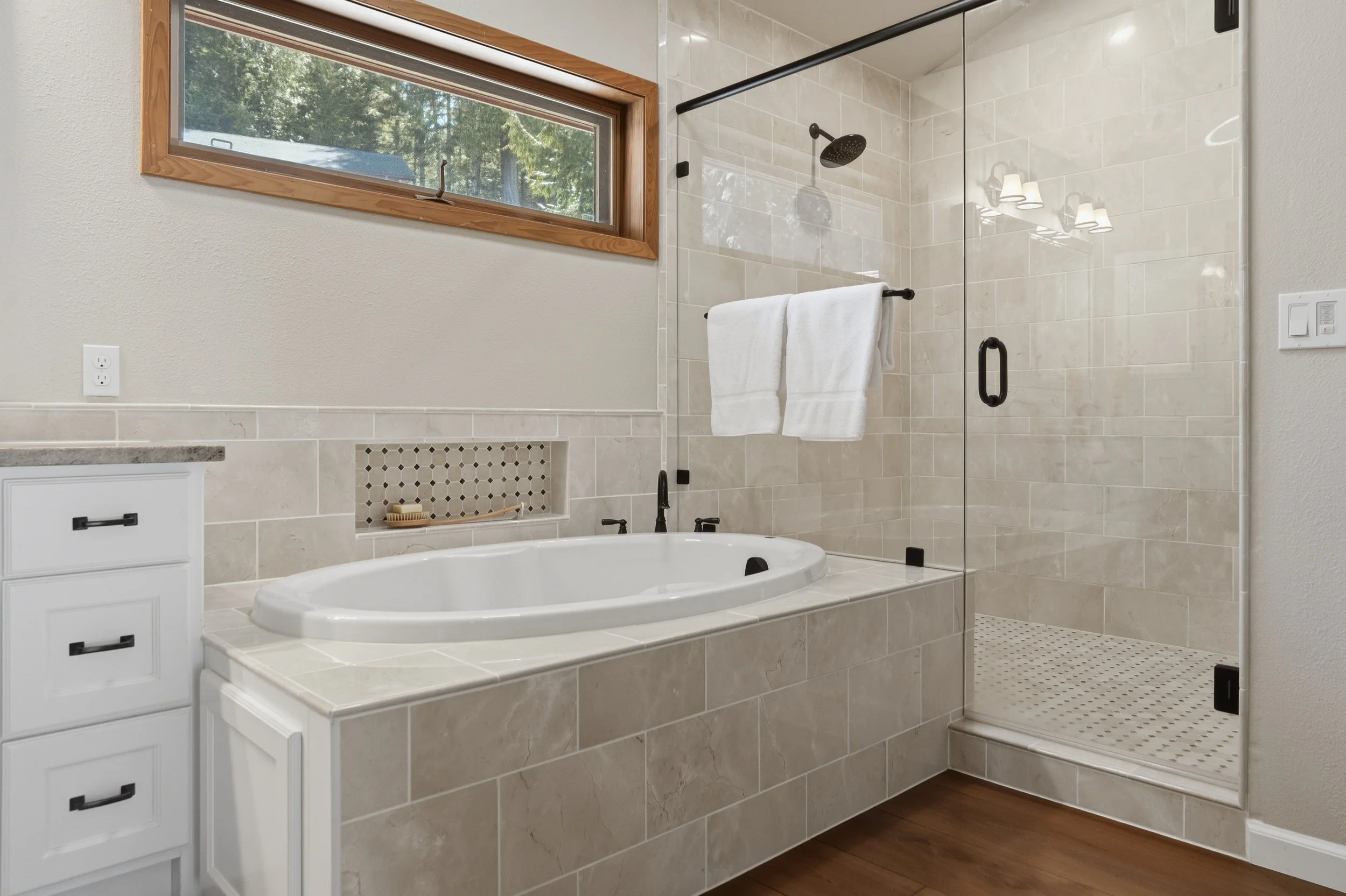 Modern bathroom with a beige tiled bathtub and a separate glass-enclosed shower area, a window with wooden trim, white cabinetry, and wood flooring.