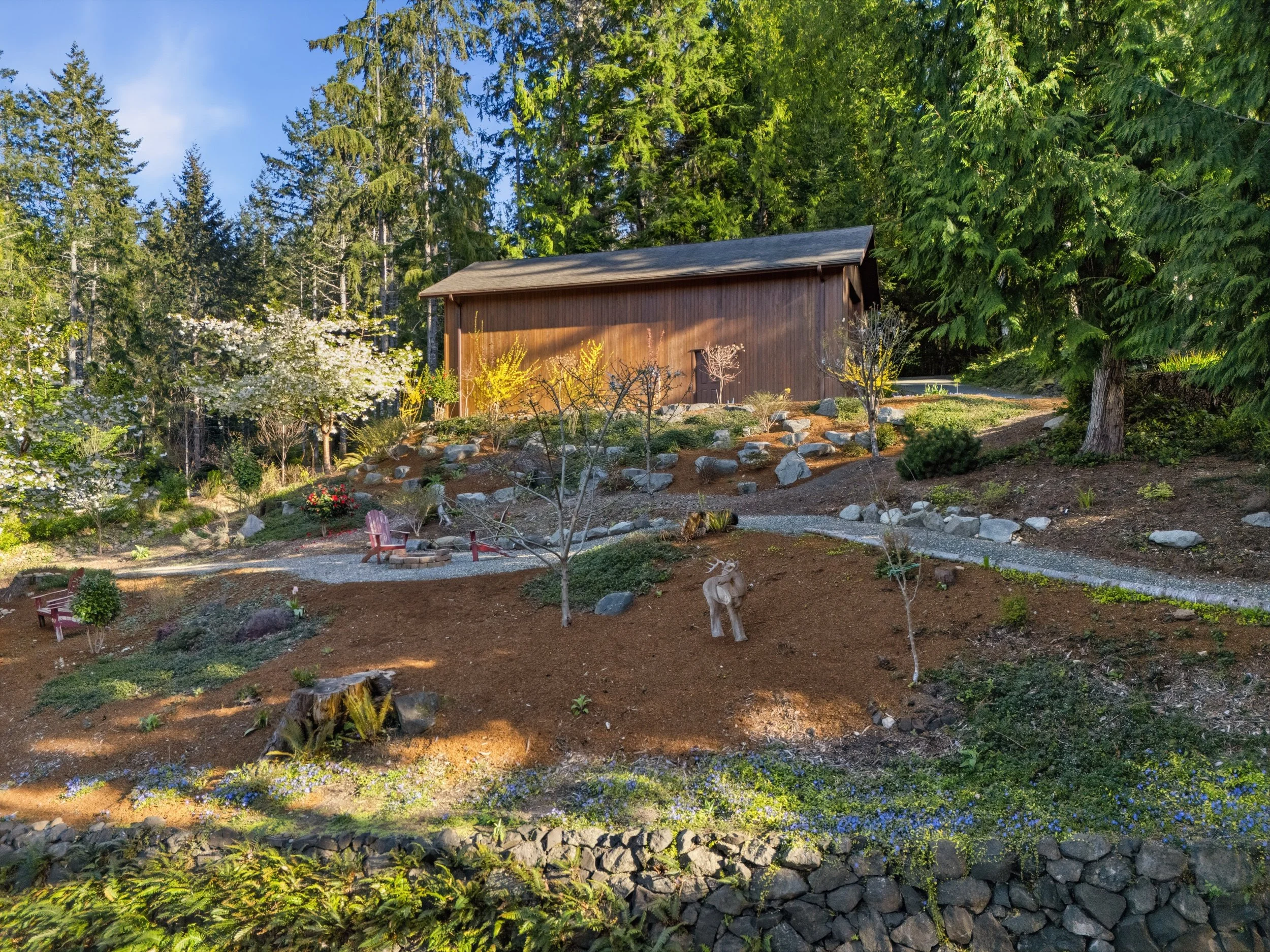 A wooden cabin situated on a hillside landscape with trees and flowers, including flowerbeds and garden decorations like a carved moose statue, around benches and a fire pit.