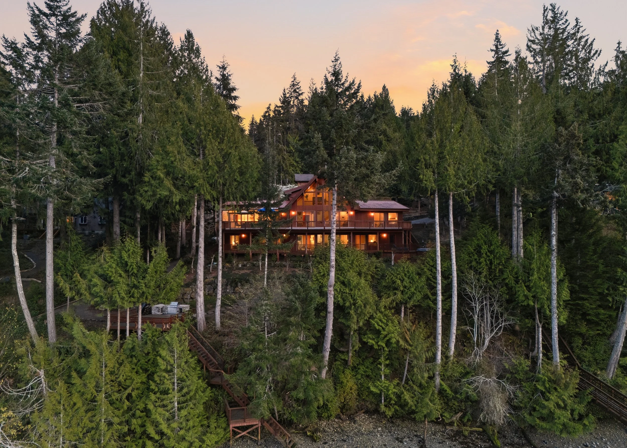 A large wooden house with multiple floors is situated on a hillside surrounded by tall pine trees, with warm lights glowing from the windows during dusk.