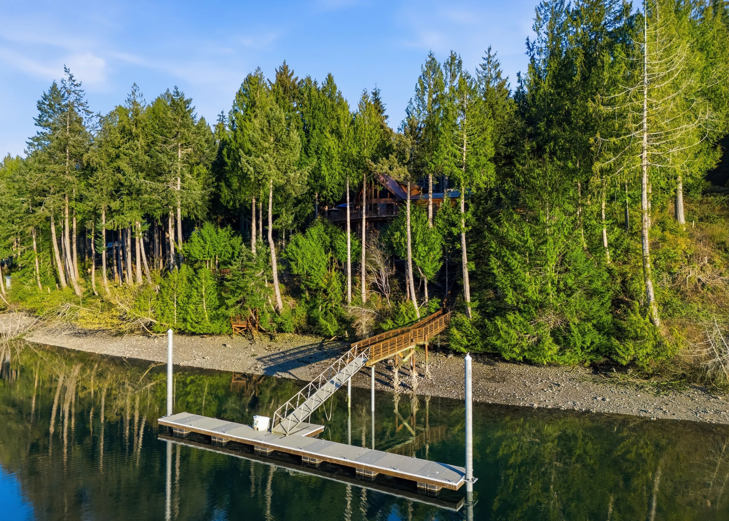 A lakeside scene with a floating dock and a ramp leading to the shoreline. The background features a house among tall green trees on a hillside, with clear blue skies overhead.