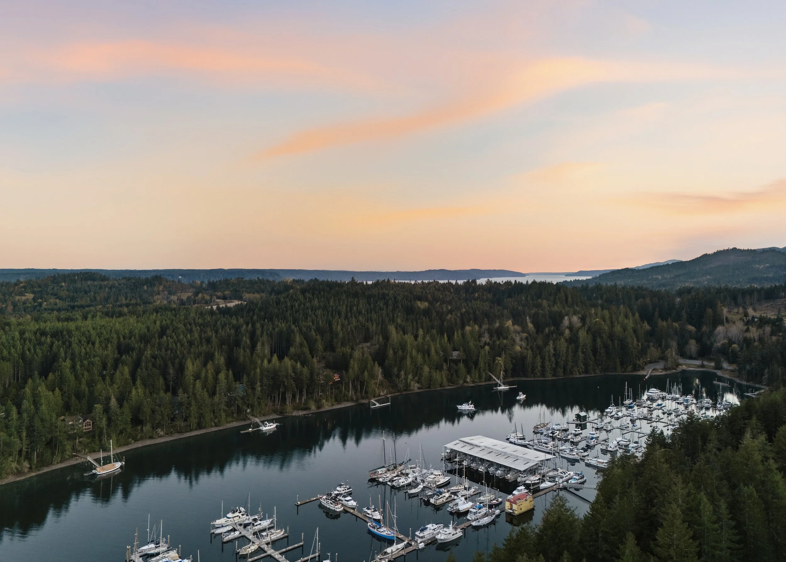 Aerial view of a marina with numerous boats docked, surrounded by dense forest and rolling hills under a pastel pink and blue sky at sunset.