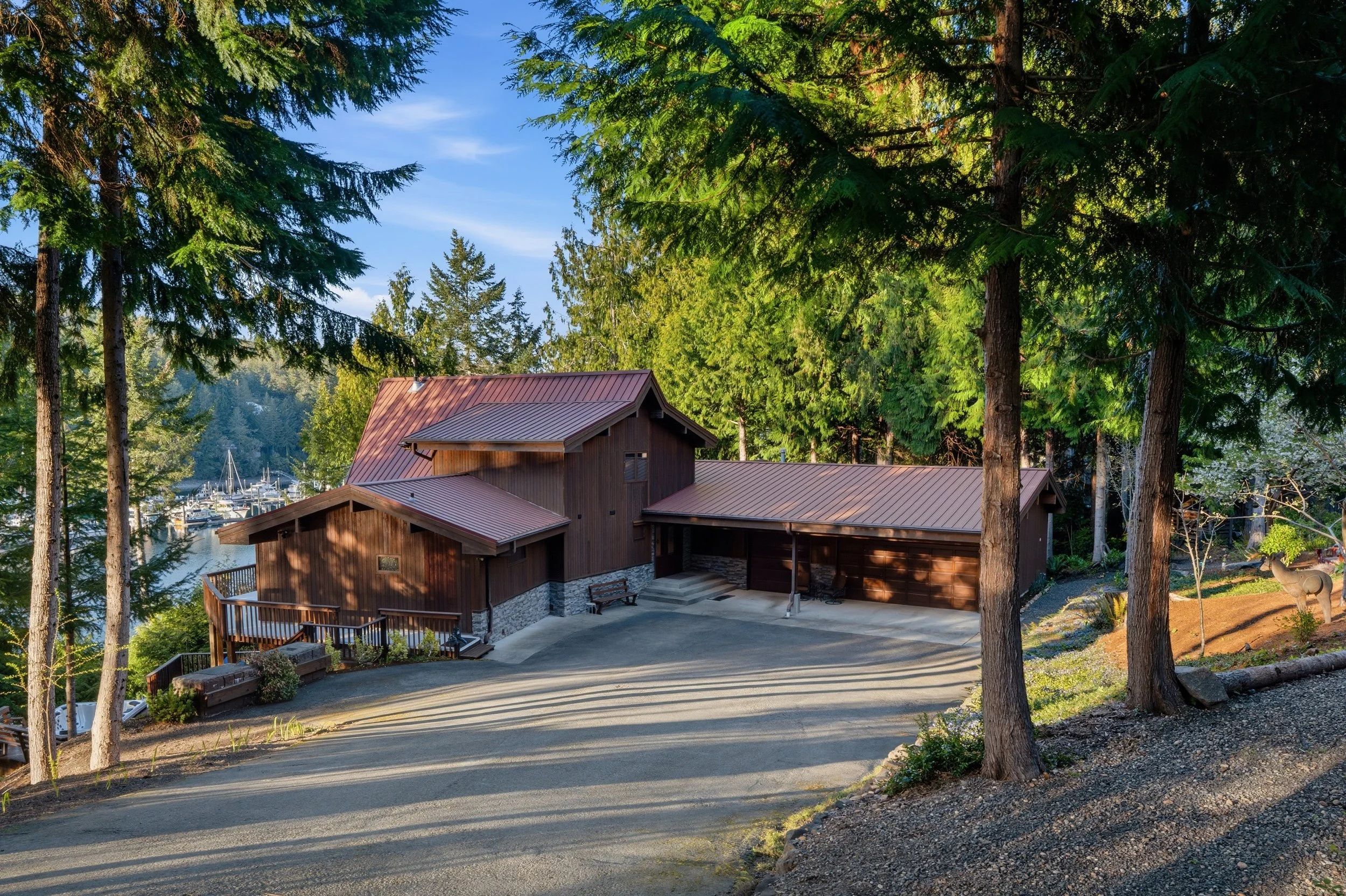 A rustic wooden house with a metal roof, surrounded by tall trees, overlooking a body of water with boats docked nearby, under a clear blue sky.