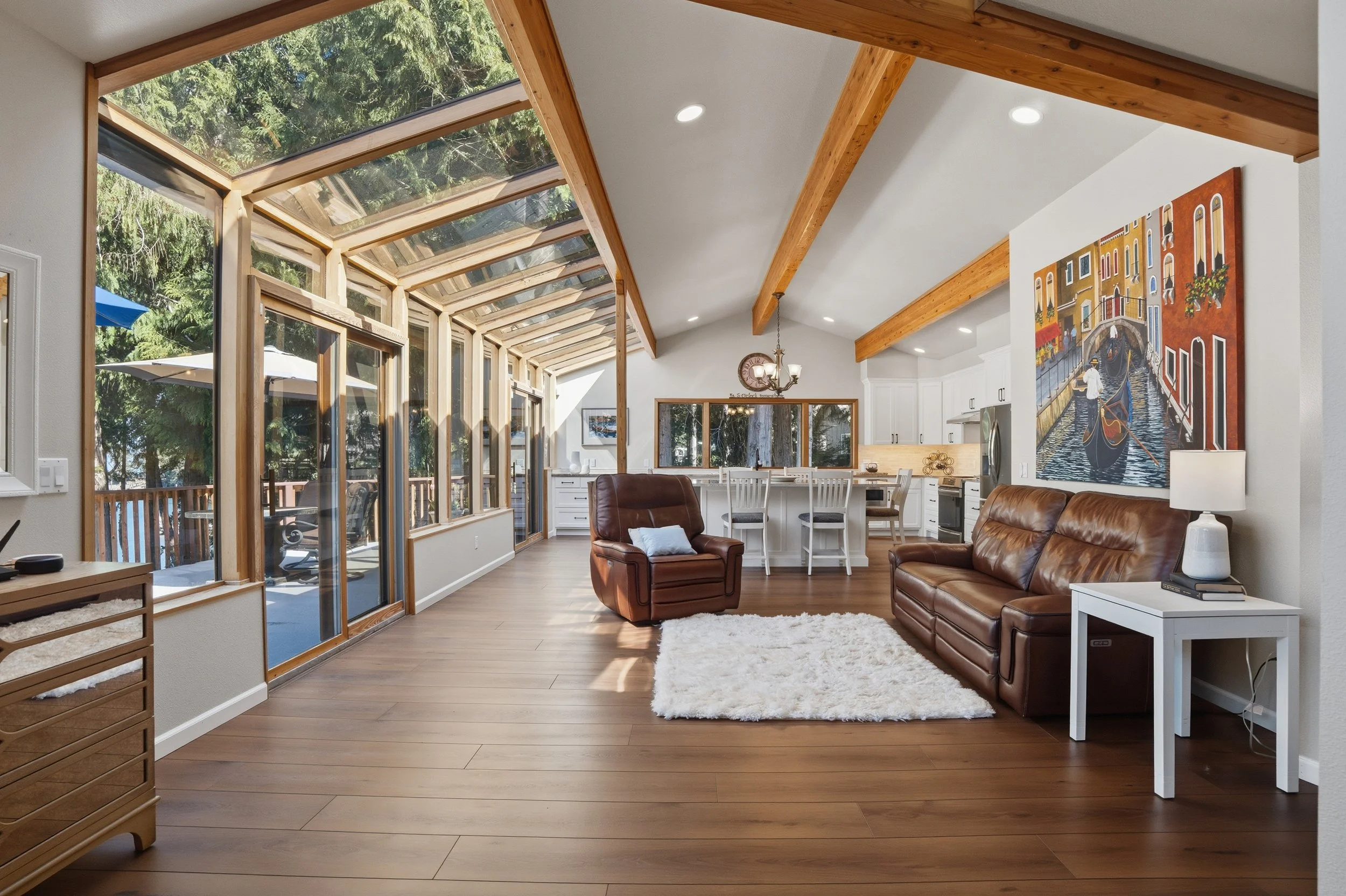Spacious open-concept living room with large sunroom featuring wooden framing, hardwood flooring, leather armchair, leather sofa, white side table with lamp, colorful artwork, kitchen with white cabinets and dining area, and large windows overlooking