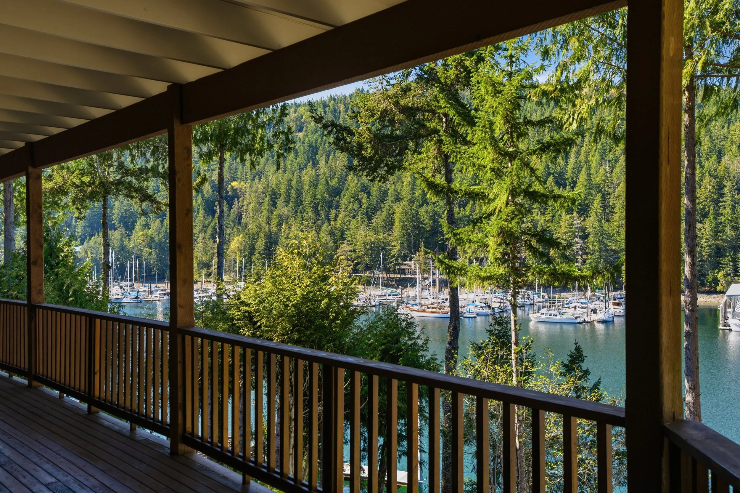 View from a wooden balcony overlooking a marina with sailboats, surrounded by dense green trees and hilly forest in the background.