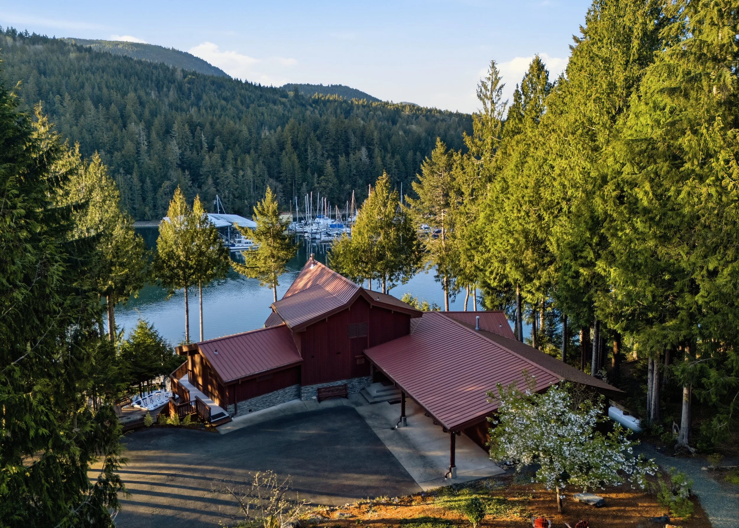 A red barn-style house surrounded by tall green trees near a lake with boats and a marina, mountains in the background.