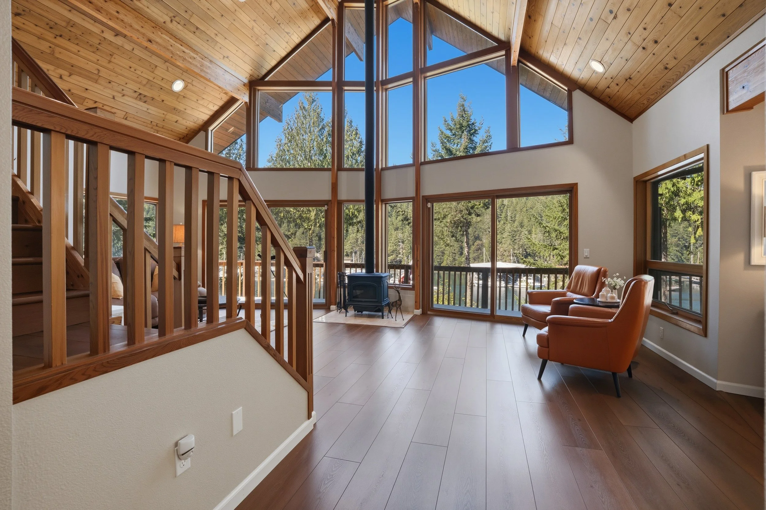Bright living room with large windows, wooden ceiling, and a wood stove, furnished with two orange armchairs and overlooks a forested yard.