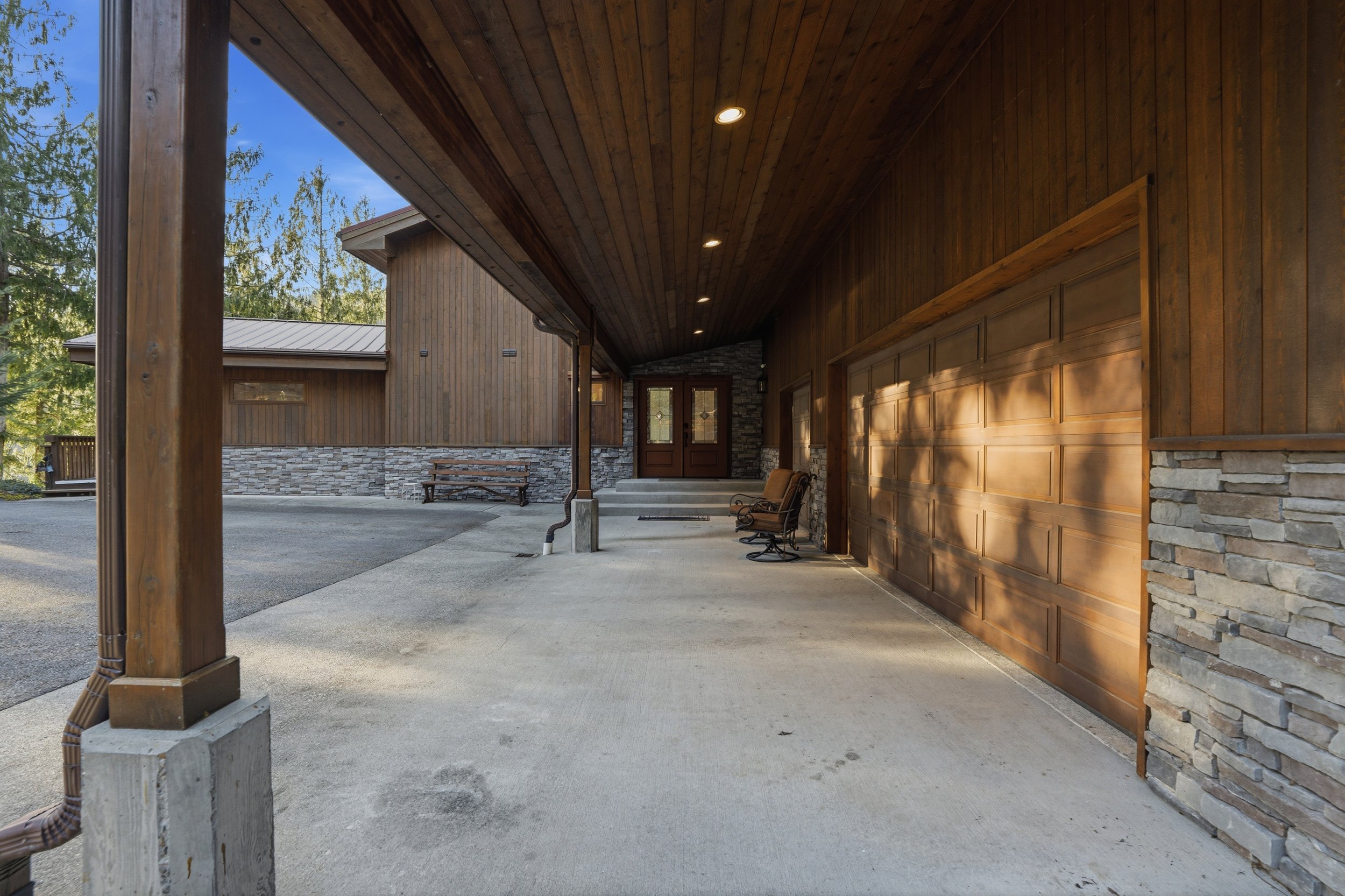 Front porch area of a house with a wooden and stone exterior, a garage door, and benches, illuminated by sunlight.