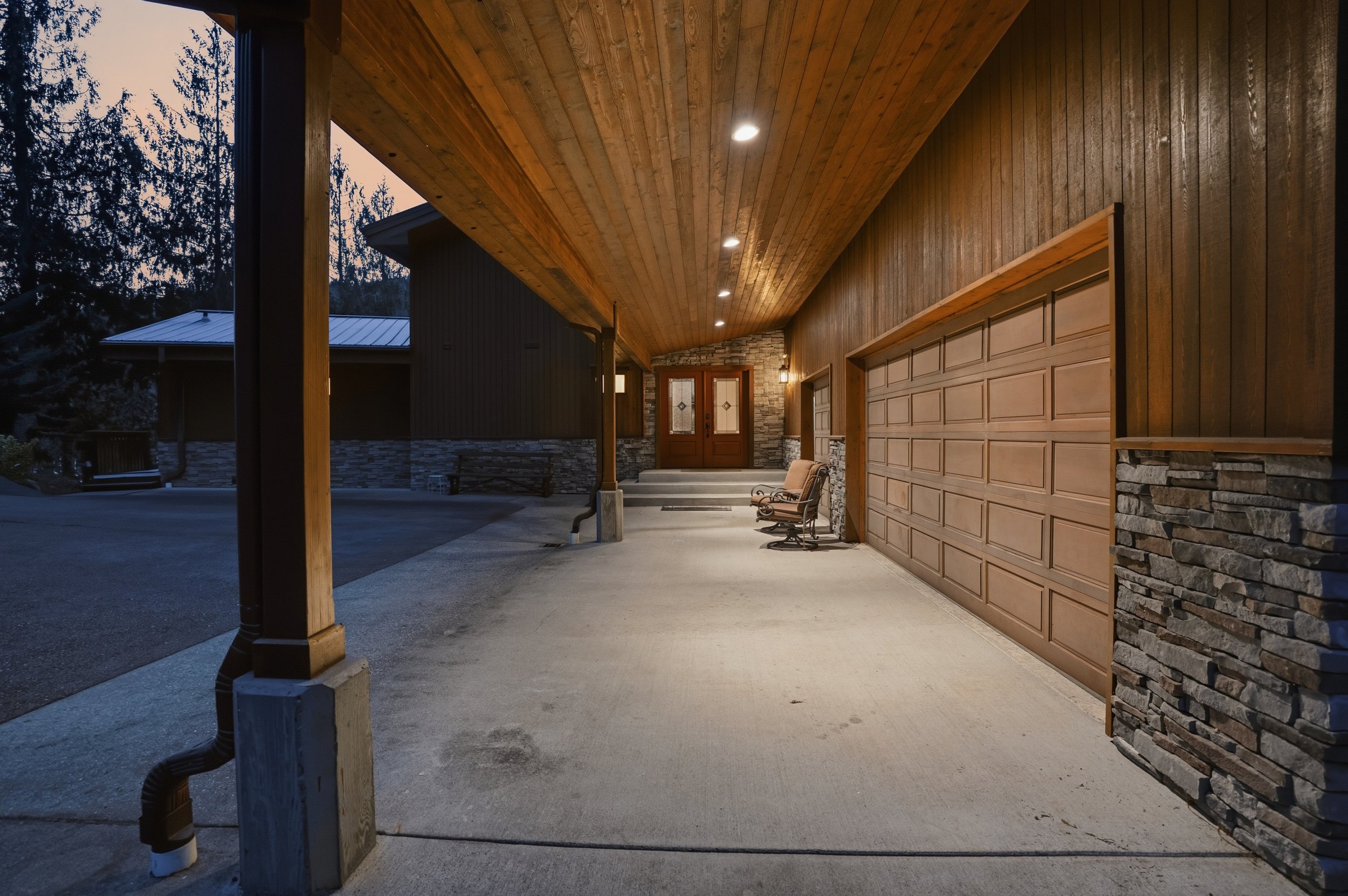 Covered driveway at dusk with wood-paneled ceiling, stone accents, and two chairs near the garage door.