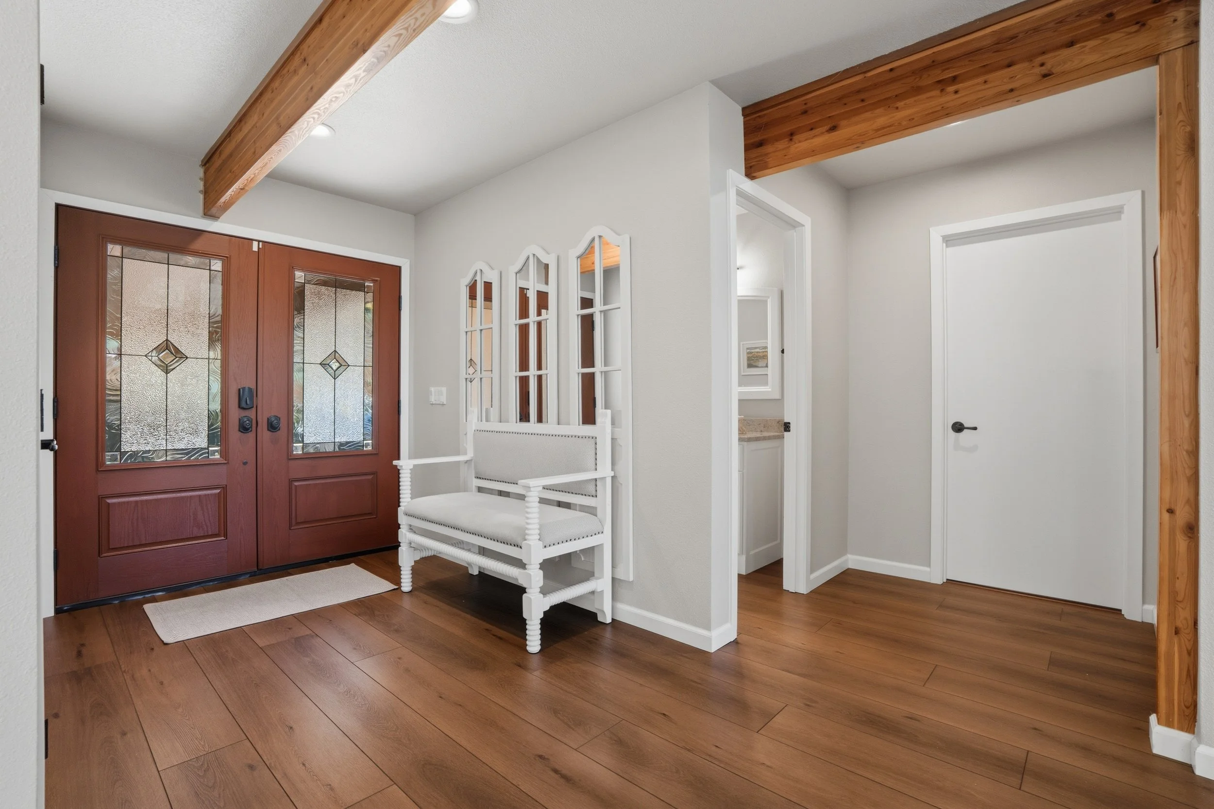 Entryway with wooden double doors, white console bench, three white decorative mirrors, and wooden flooring.