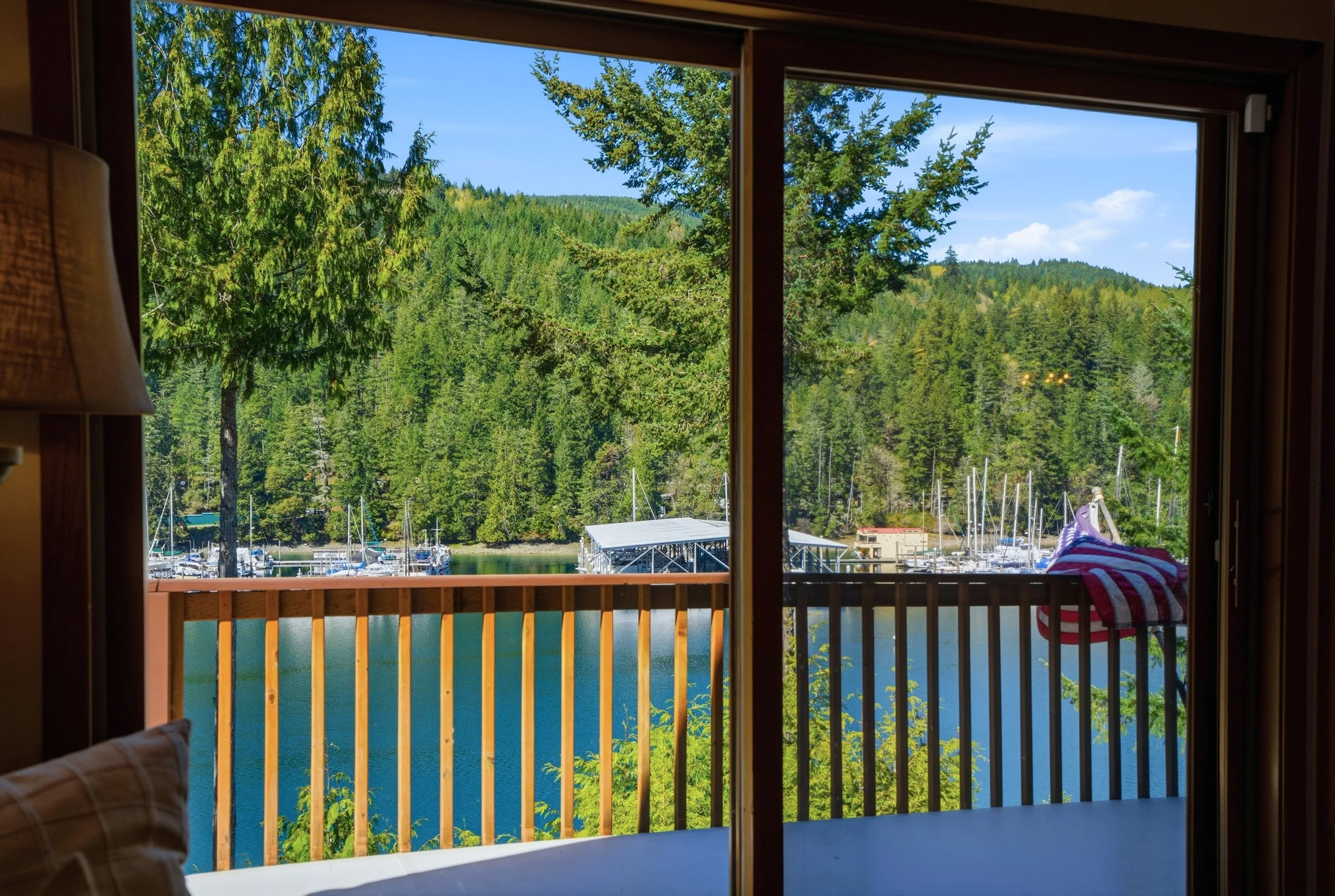 View of a lake with boats and a dock, surrounded by a forested hillside, seen through a sliding glass door with a wooden railing and an American flag on the balcony.