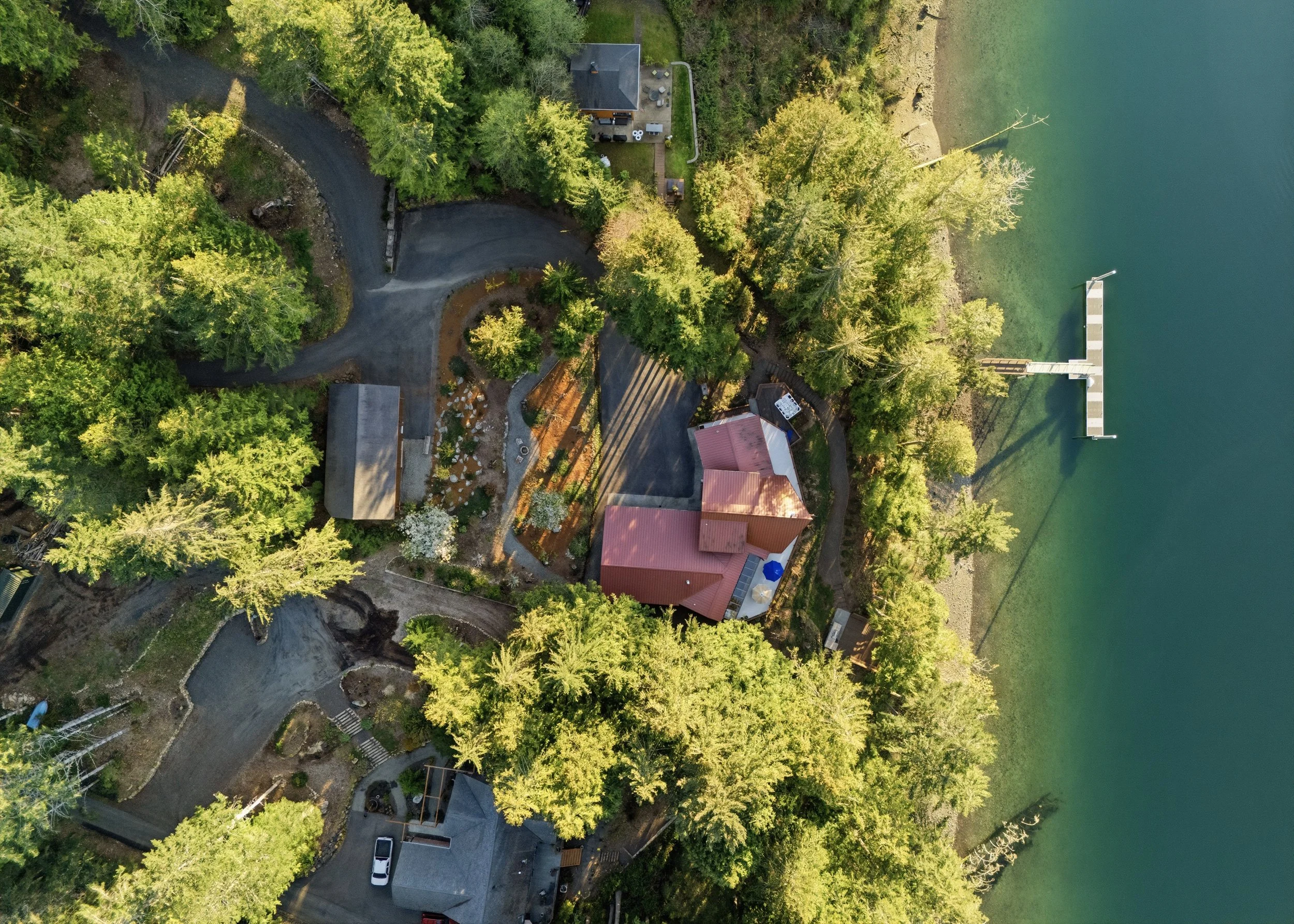 Aerial view of a lakeside property surrounded by trees, featuring a house with a red roof, a dock extending into the water, and a winding driveway.