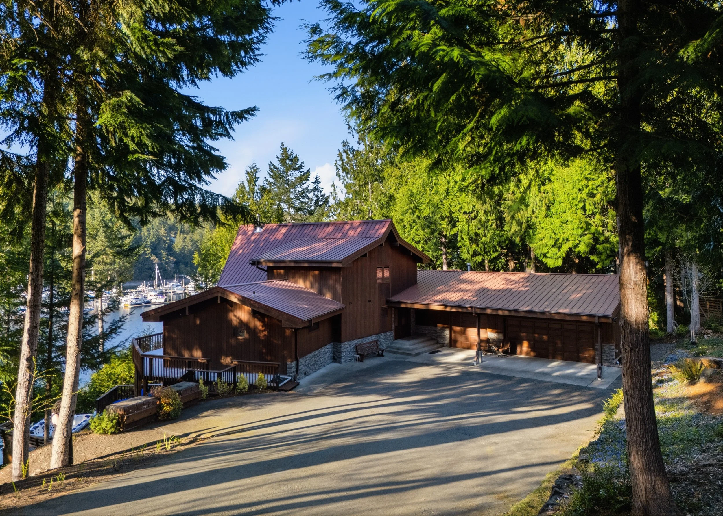 A wooden house with a metal roof situated among tall green trees, with a garage and a concrete driveway. In the background, there are boats docked at a marina on a body of water, under a blue sky.
