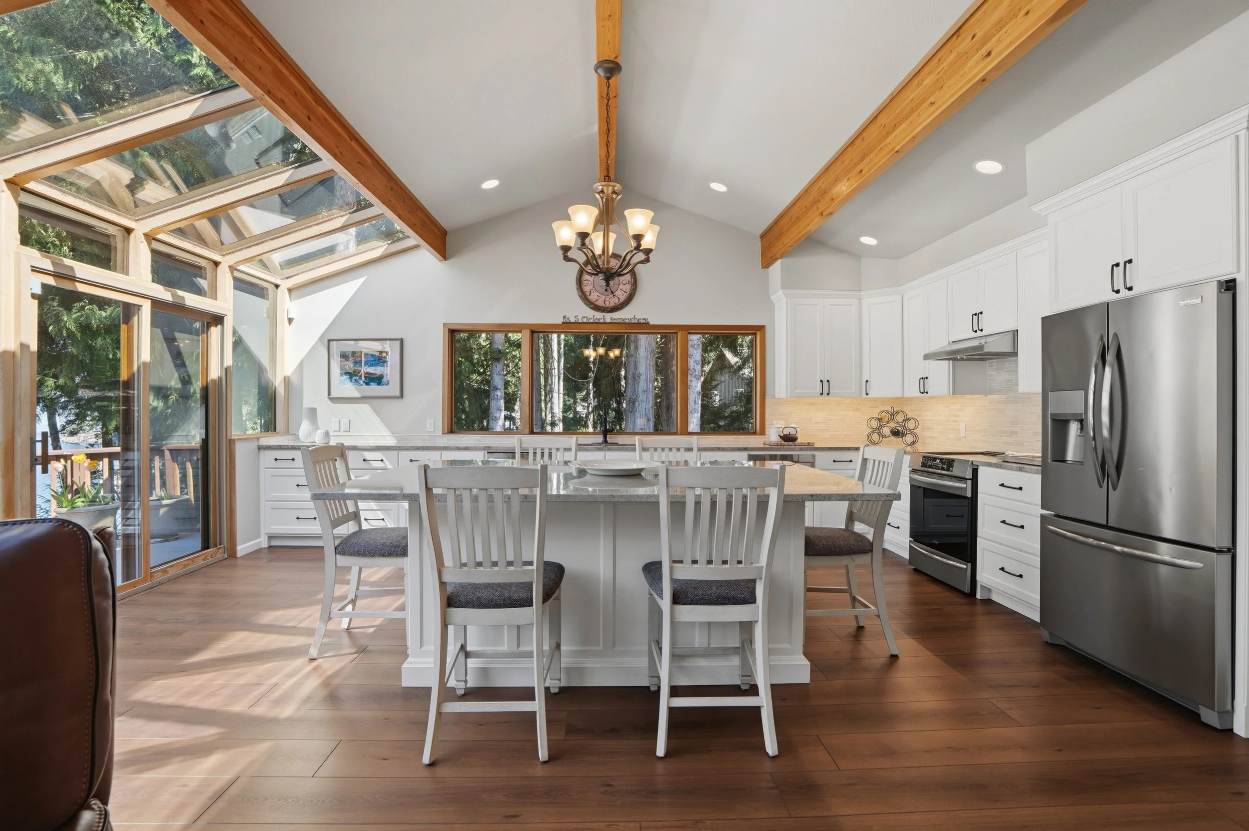 Bright kitchen with white cabinets, stainless steel refrigerator, black appliances, wooden beams and high vaulted ceiling with skylights, central island with seating, and large window with view of trees.