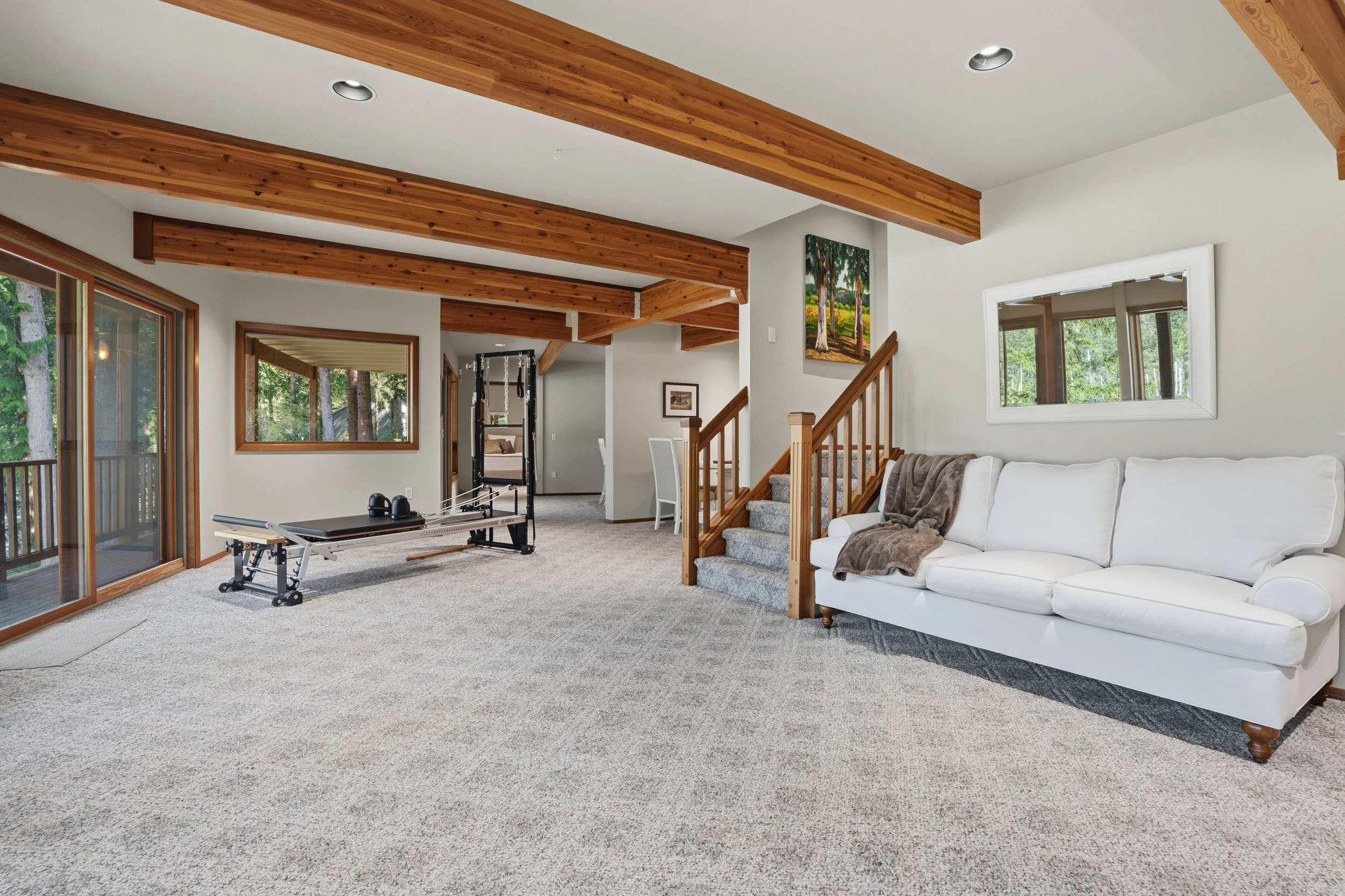 Living room with beige carpet, white sofa with brown blanket, wooden staircase, sliding glass doors, and windows showing trees outside.