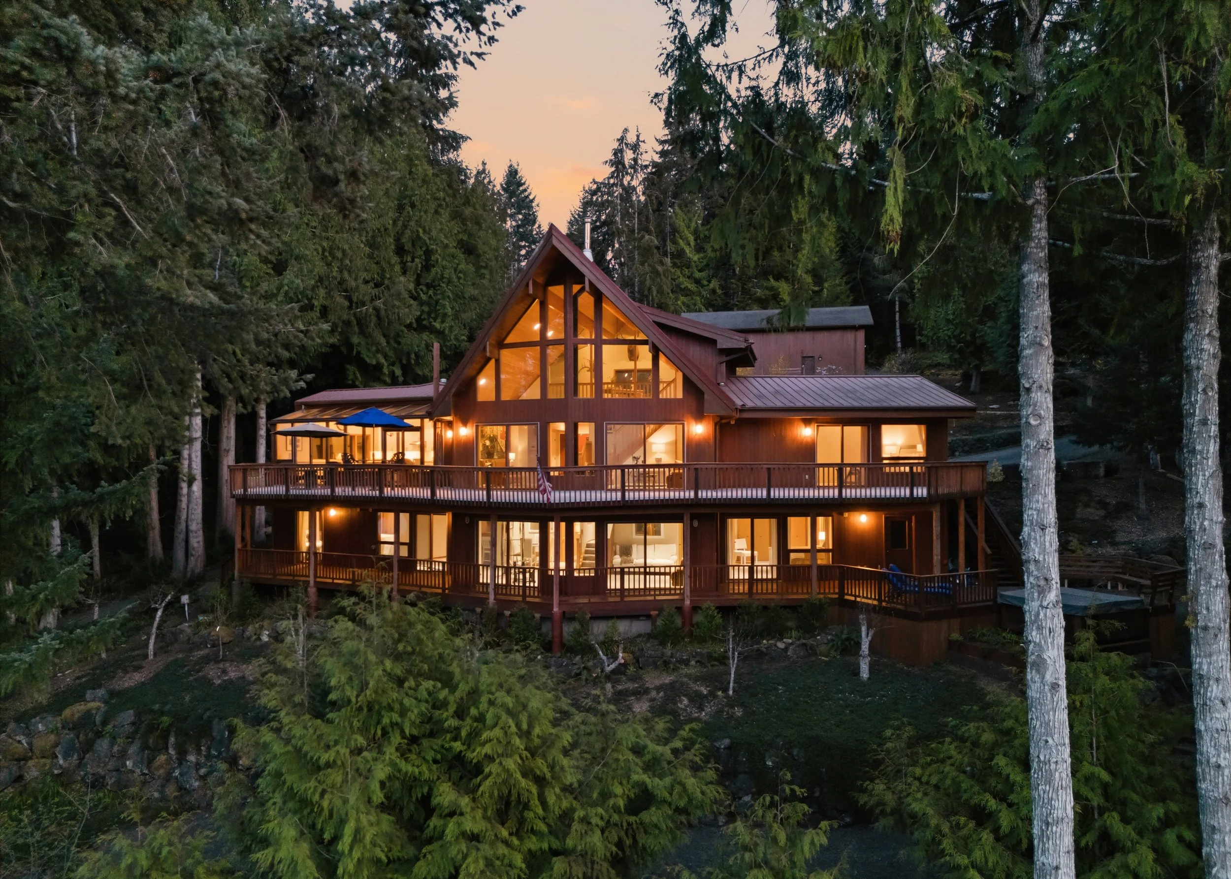 A large house with multiple stories and a prominent A-frame roof is situated among tall trees, illuminated from within during dusk, with an expansive deck wrapping around it.
