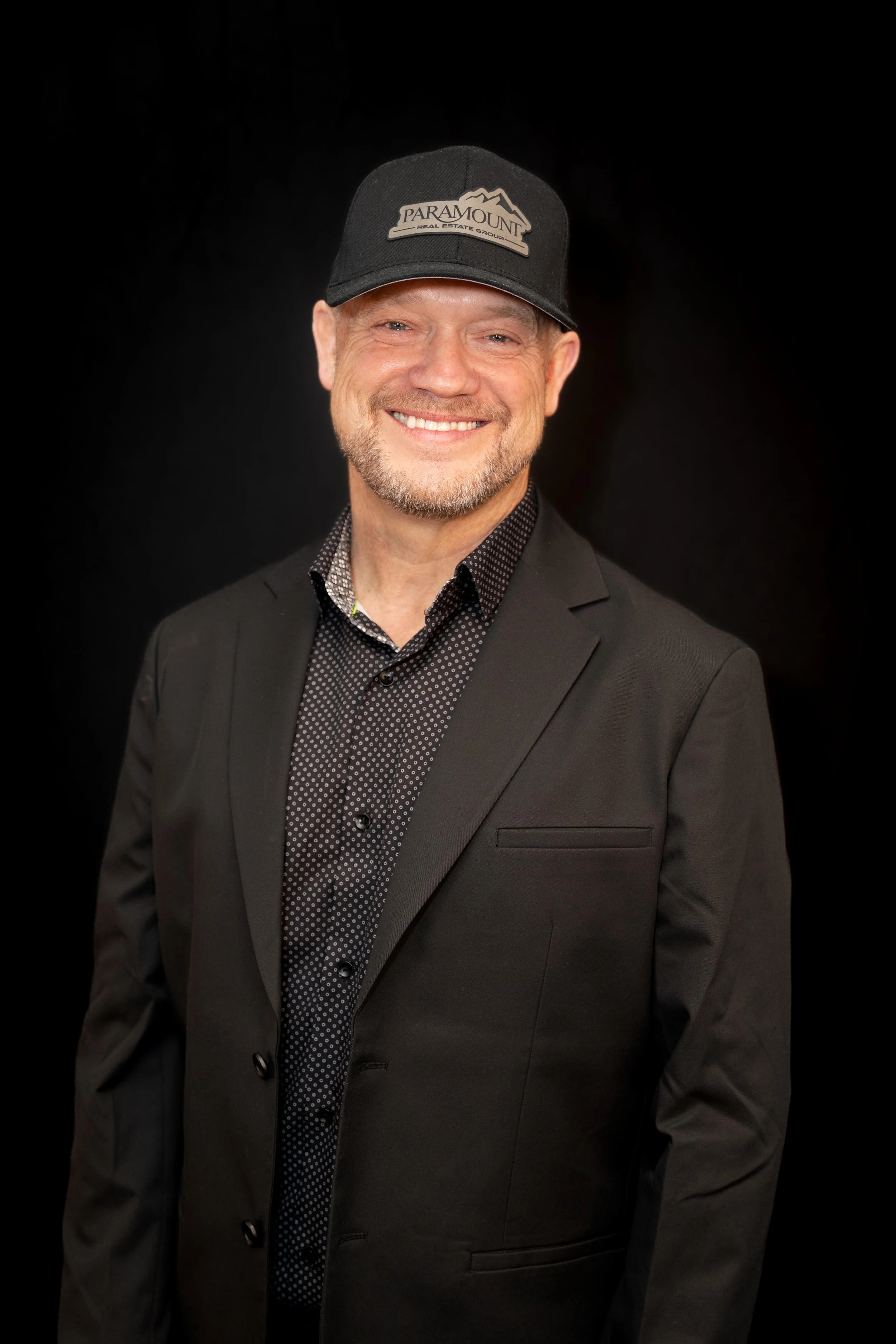 A man smiling, wearing a black blazer and a black baseball cap that says 'Paramount Real Estate Group' against a black background.