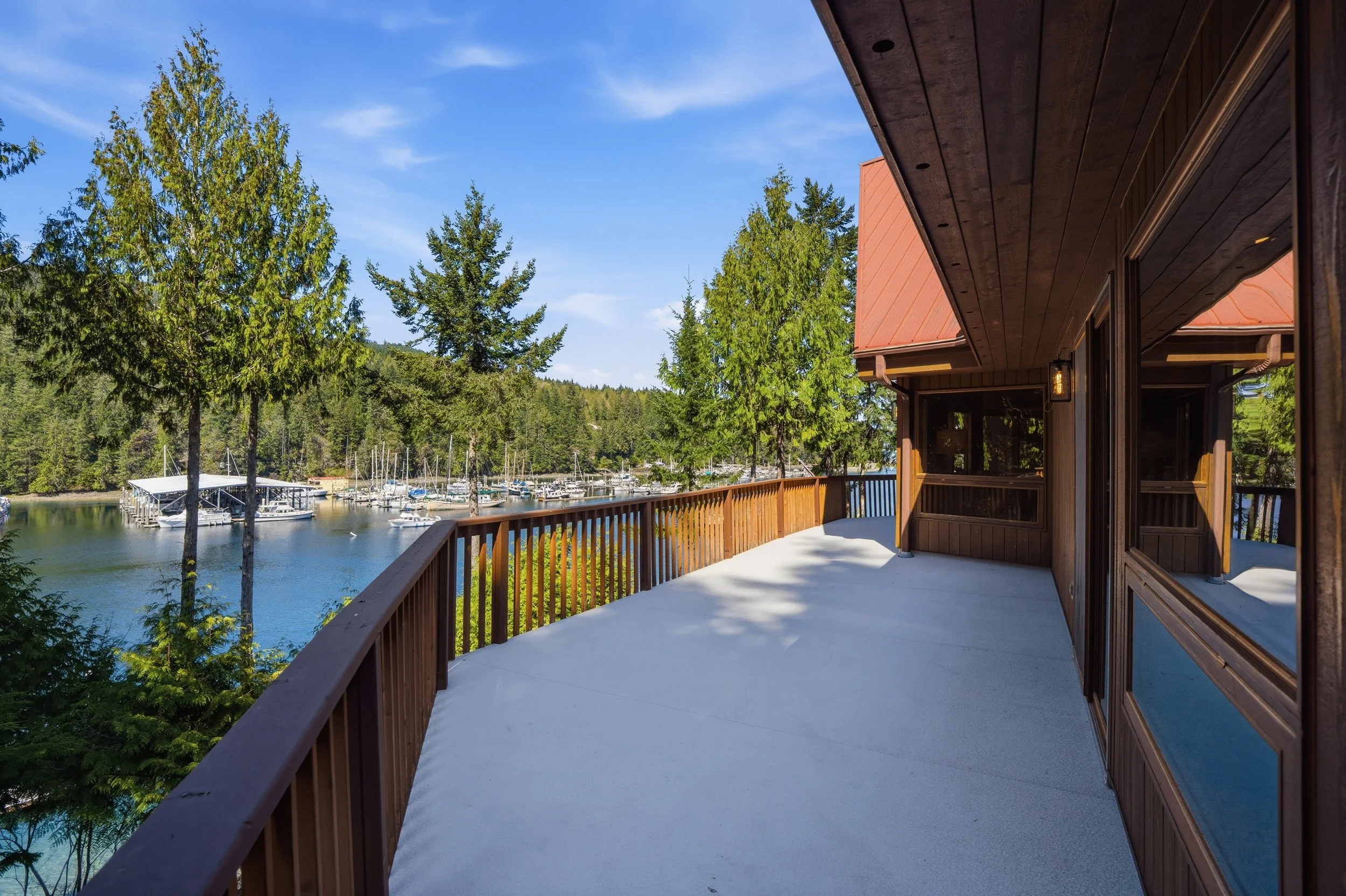 View of a balcony overlooking a marina with sailboats, surrounded by tall green trees and a clear blue sky.