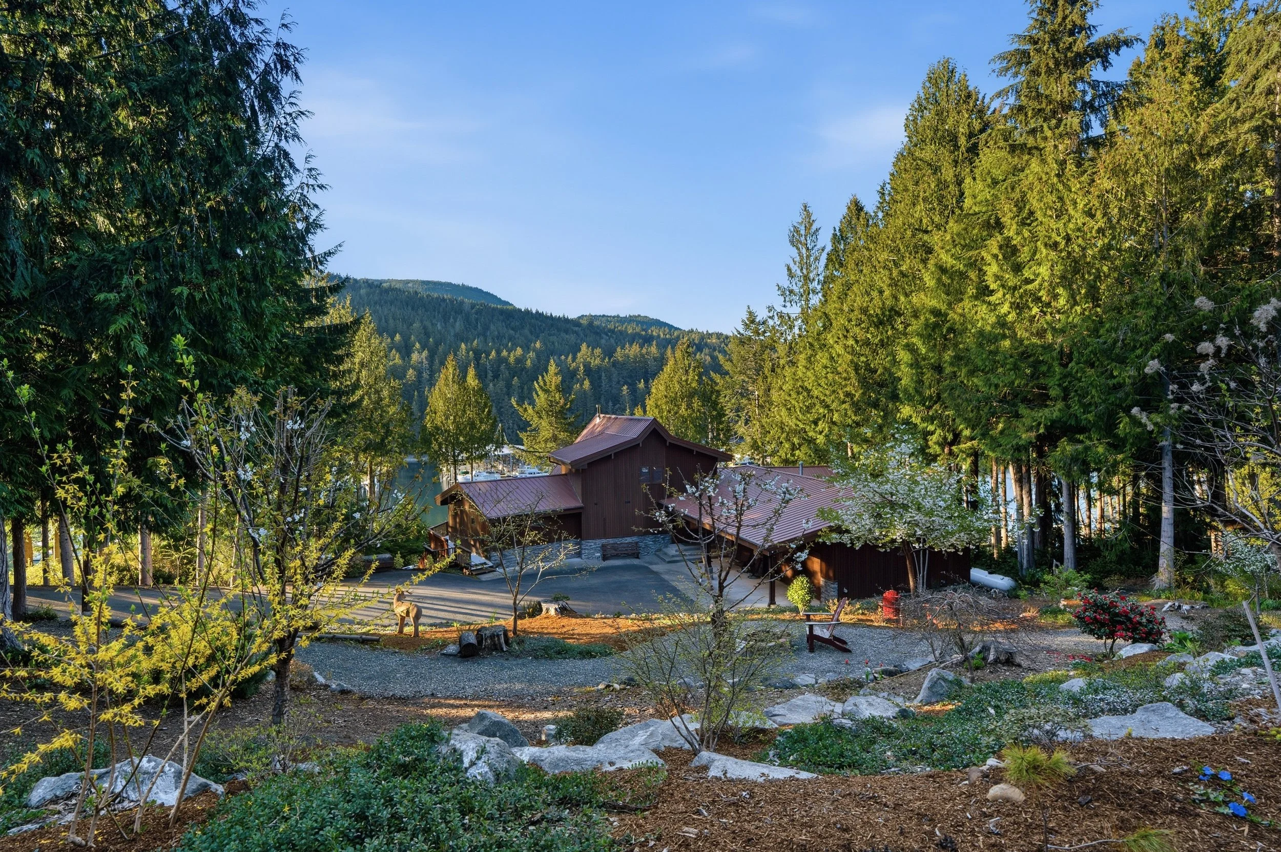A scenic landscape of a forested area with a wooden house and a lake in the background, surrounded by tall evergreen trees and mountains.