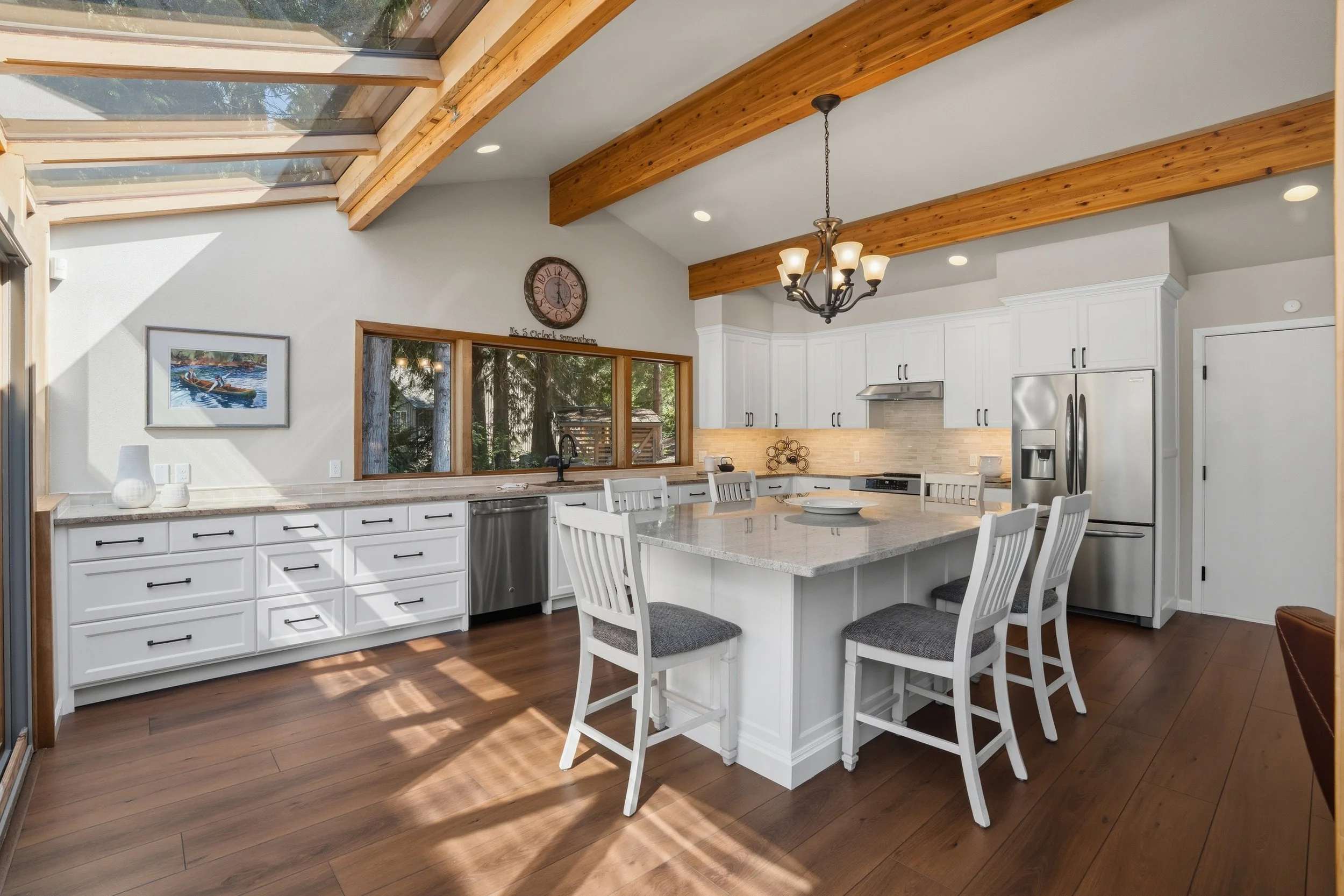 Modern kitchen with white cabinetry, a large island with seating, stainless steel appliances, and wooden beams on the ceiling. Large windows and a skylight let in natural light.