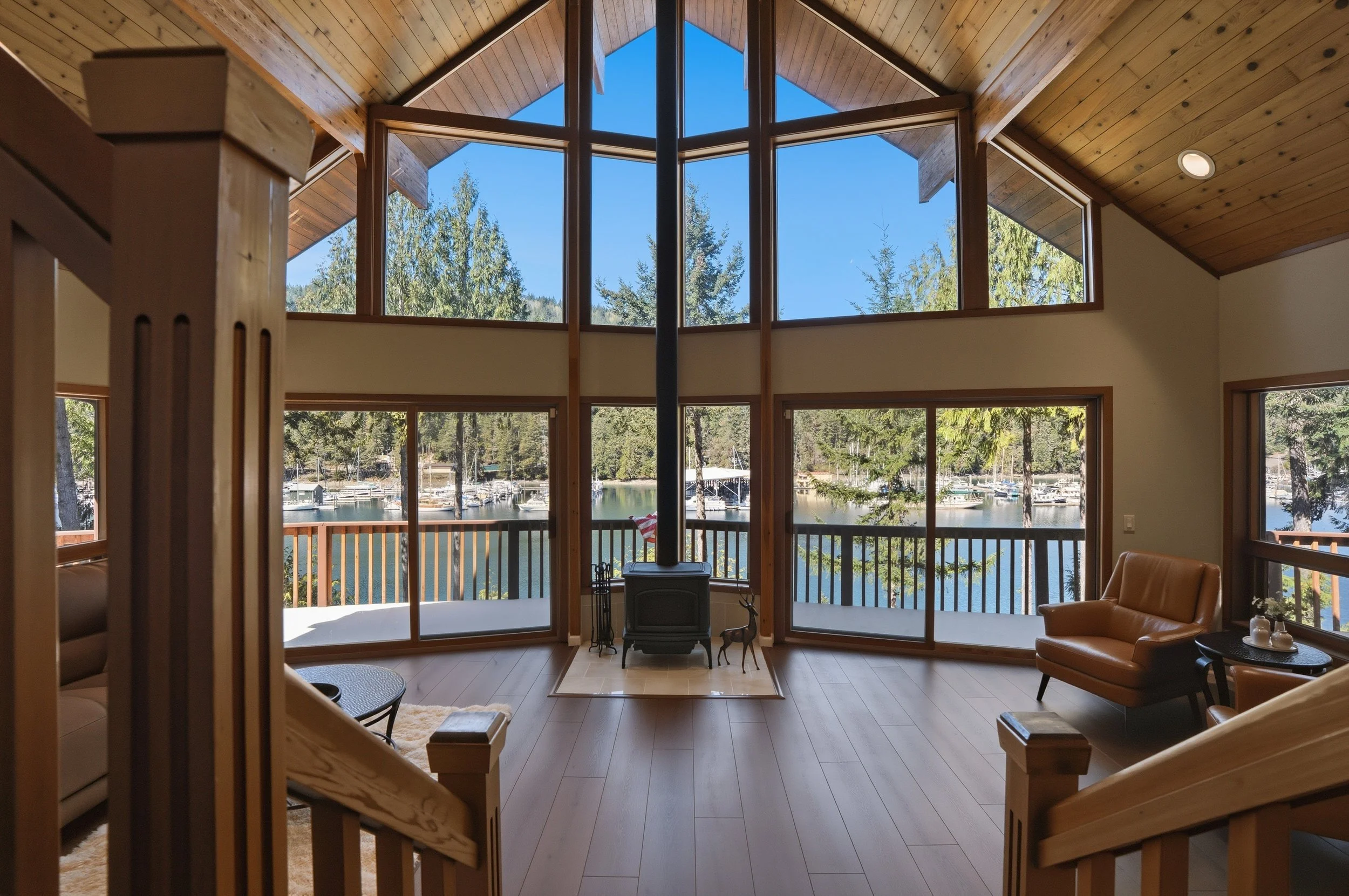 Living room with large windows and a view of a lake with boats, trees, and a clear blue sky. Wood stove at the center, with seating and a railing on the deck outside.
