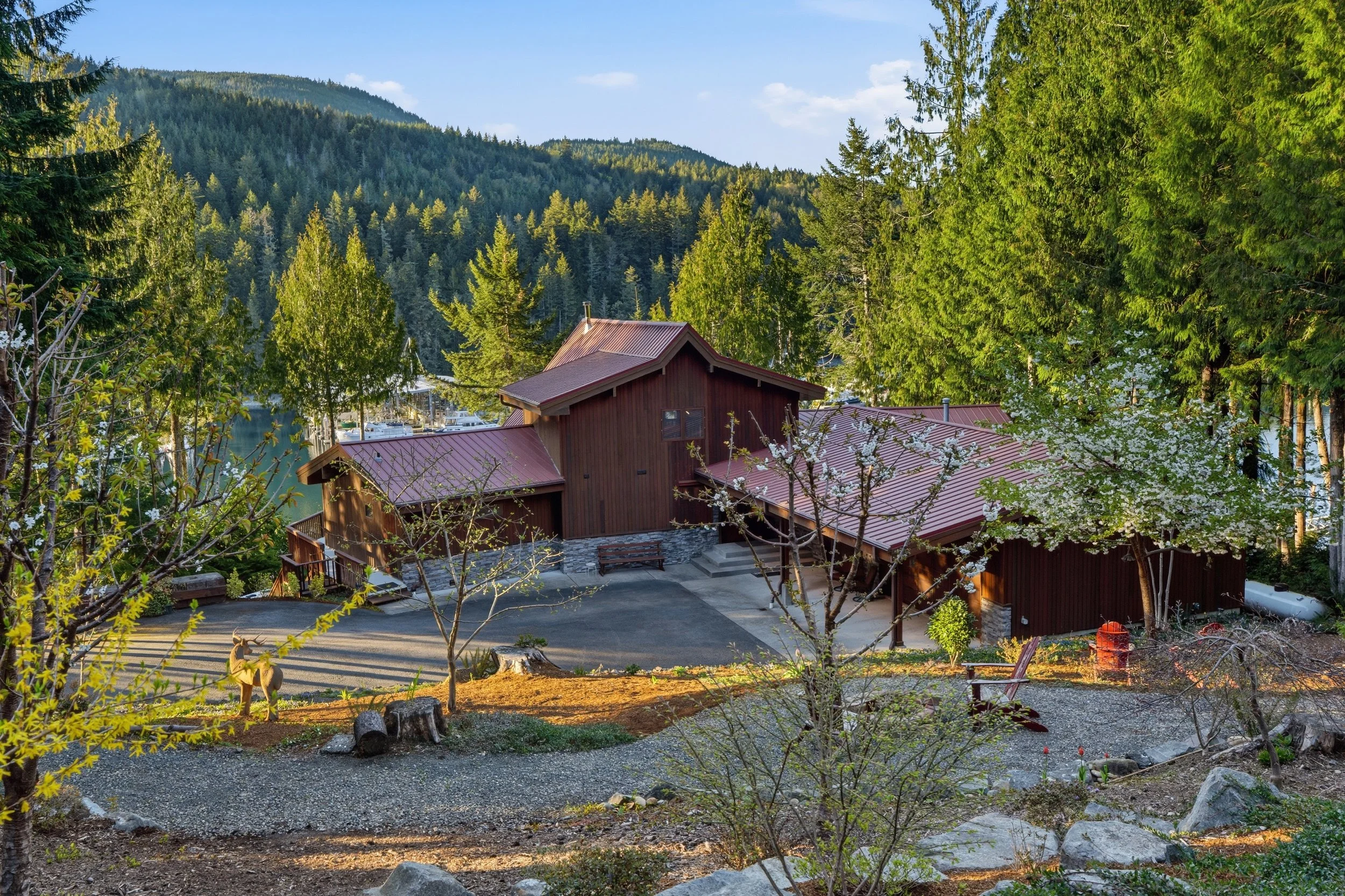 A large wooden house with a red roof is surrounded by trees and mountains, with a gravel pathway and flowers in the yard.