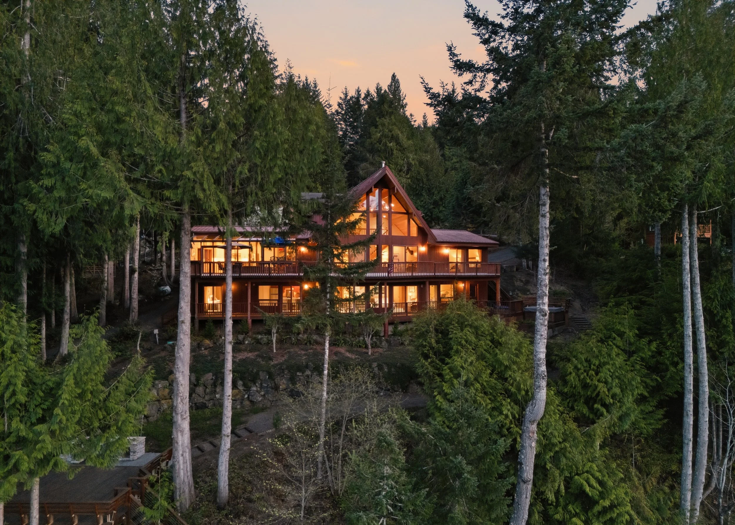 A large house with multiple levels and extensive windows, illuminated from within, surrounded by a dense forest of tall trees at dusk.