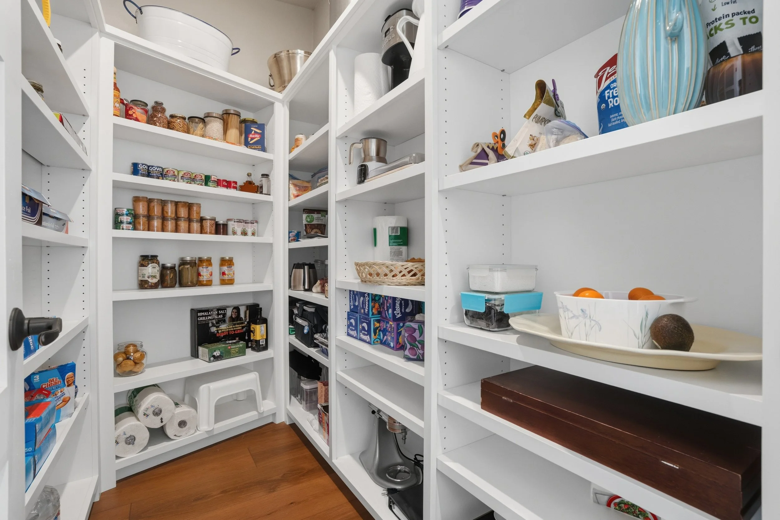 White pantry shelves filled with food items, jars, containers, and kitchen appliances, with some paper towels and miscellaneous kitchen tools visible.