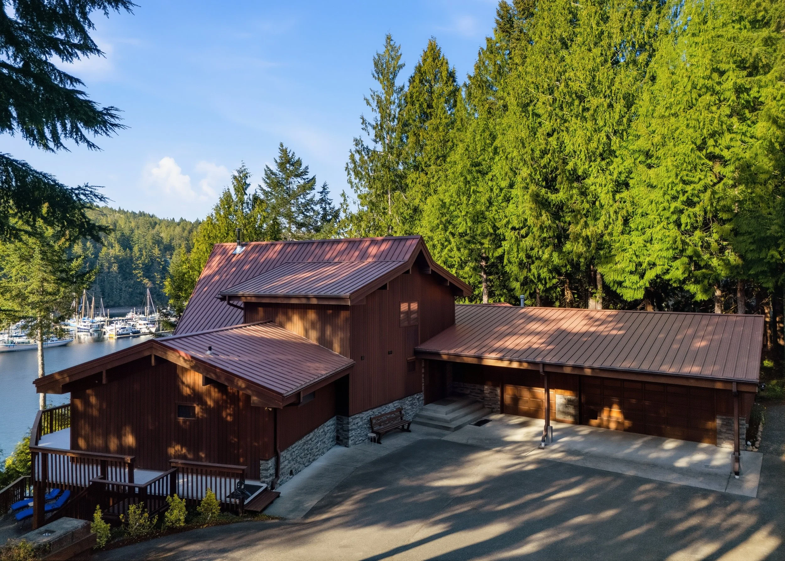 A large wooden house with a metal roof situated near a forest and a marina with boats on a body of water.