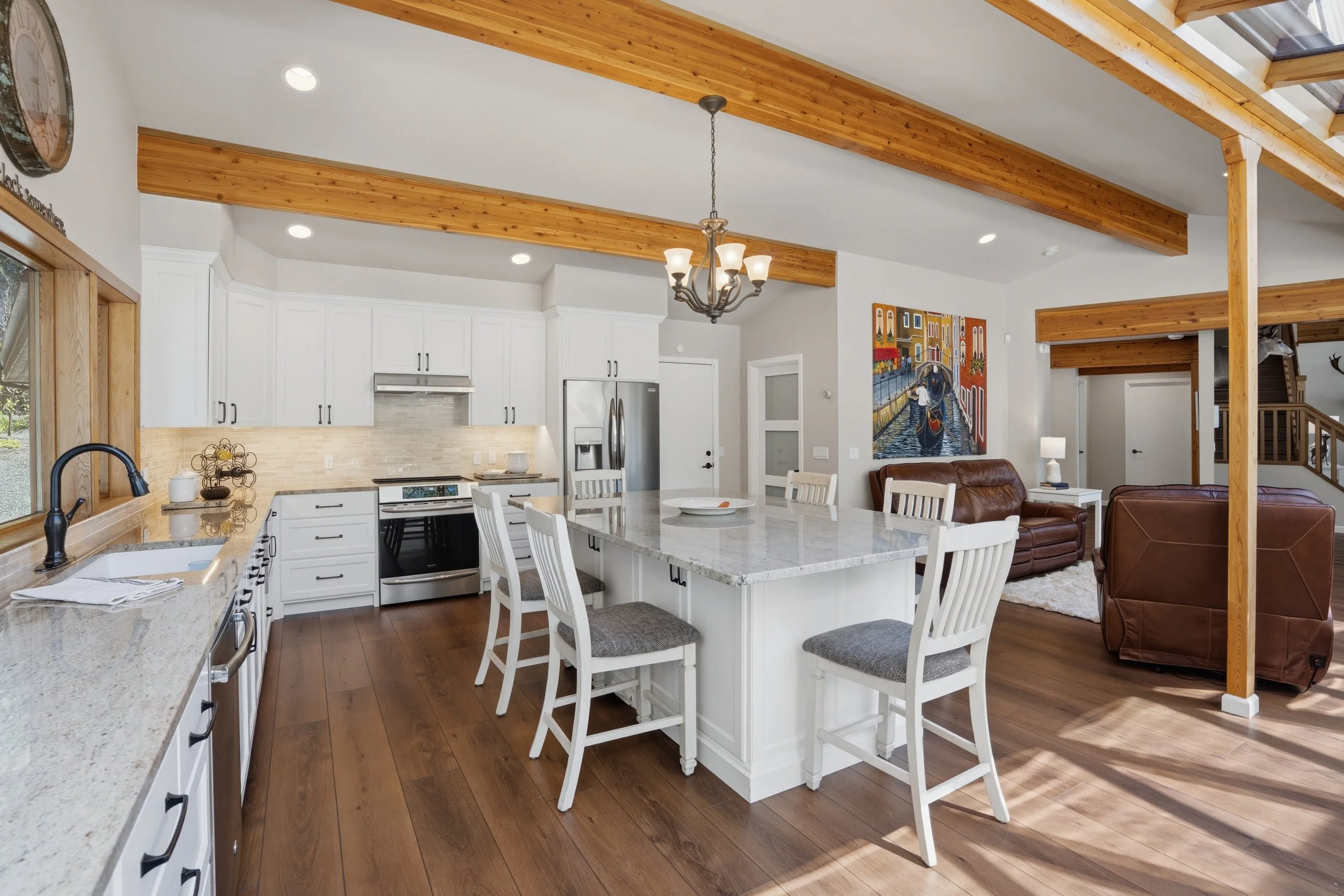 Open concept kitchen and living room with white cabinets, stainless steel appliances, granite countertops, a kitchen island with white chairs, wooden beams on the ceiling, hardwood flooring, and wall art in the living area.
